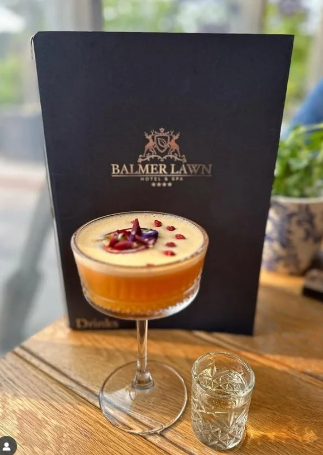 A cocktail drink with a yellow-orange color in a glass with a wide bowl and a stem, garnished with red petals or pomegranate seeds and purple flower petals. A small glass of clear liquid, possibly water, is next to it on a wooden table. A menu or promotional card for Balmer Lawn Hotel & Spa is in the background.