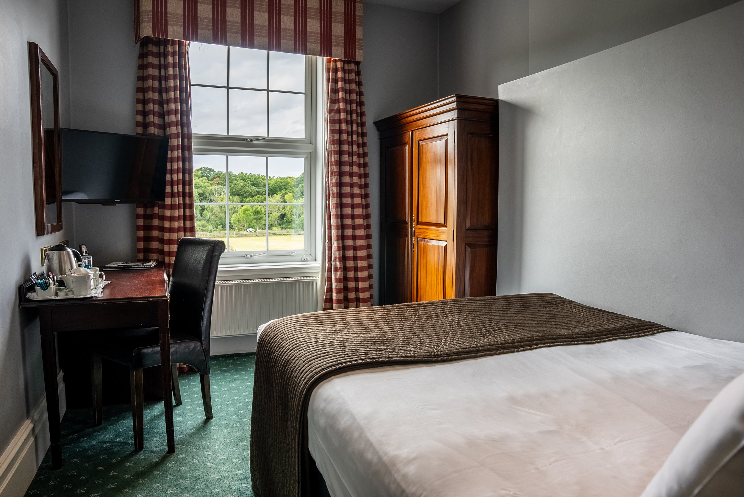 Hotel room with a bed, wooden wardrobe, window with checkered curtains, desk with a chair, and a small TV mounted on the wall.