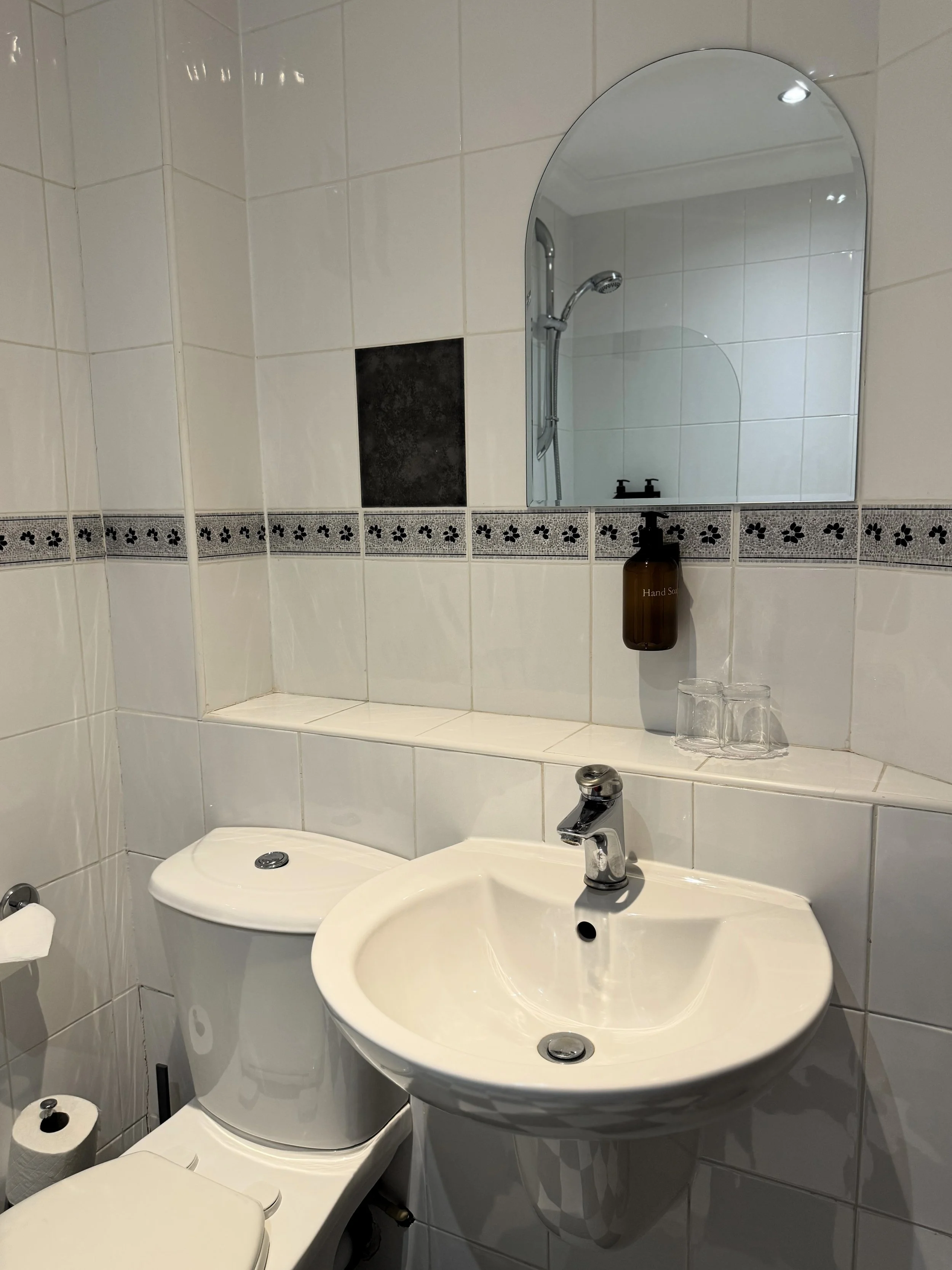 A small bathroom with white tiled walls, a round mirror above a small white sink, a brown hand soap dispenser, and two glasses on a tiled ledge. Part of a white toilet and a toilet paper roll are visible. A shower area with a curved glass door and a 
