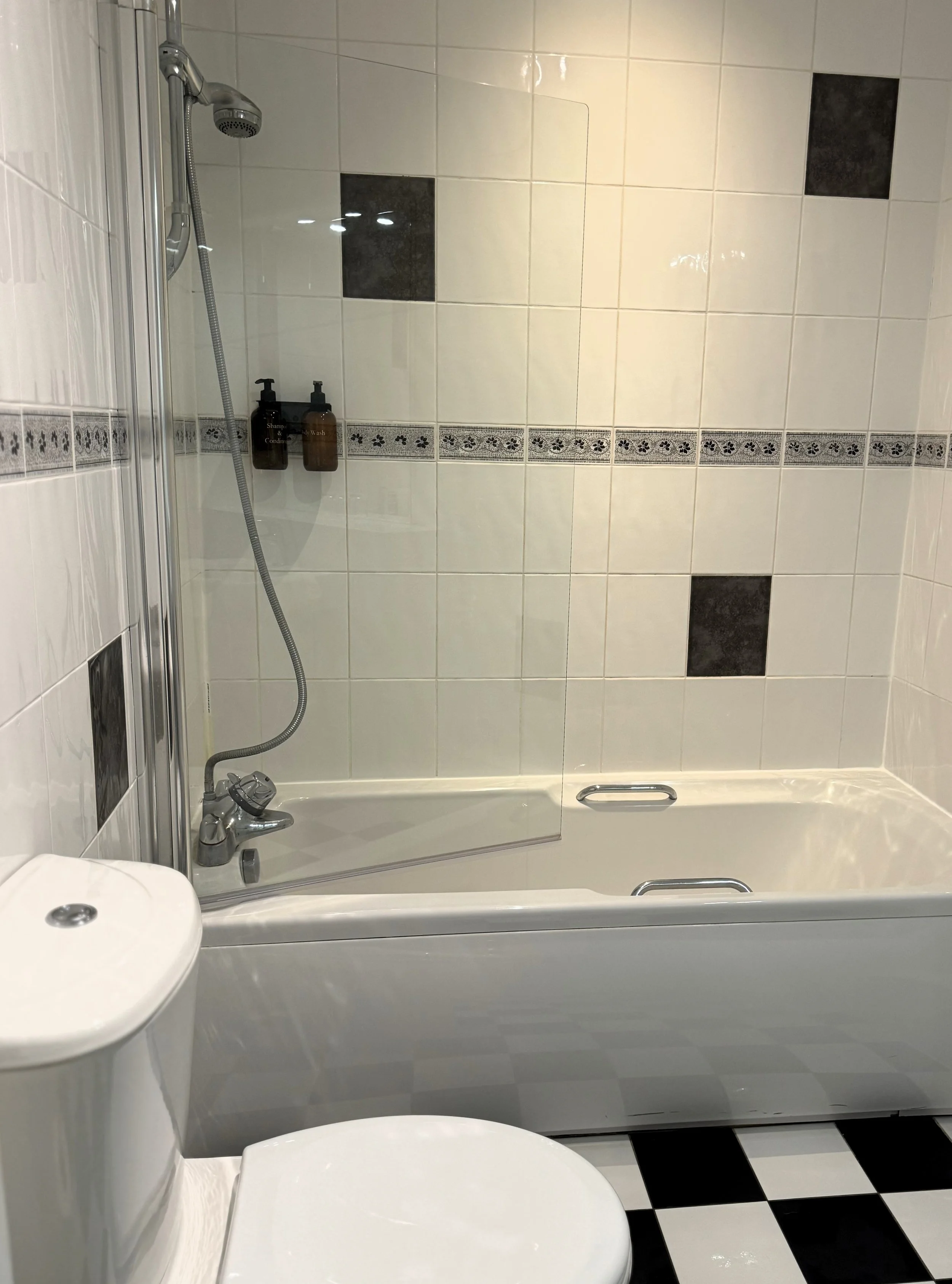 Bathroom with a white toilet, bathtub with a glass shower door, and a tiled wall with black and white patterns.