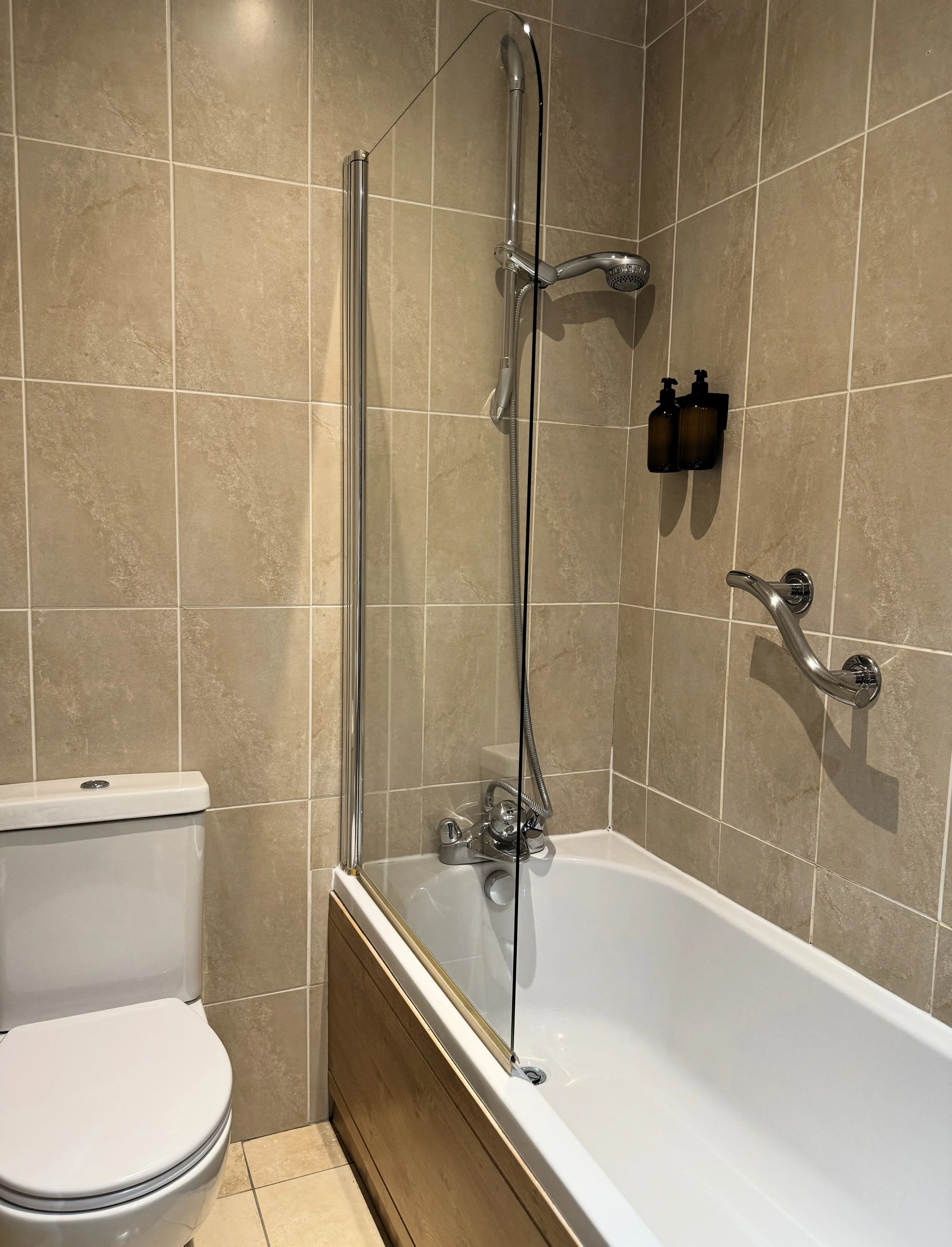Bathroom with a bathtub/shower combination, beige tiled walls, a handheld showerhead, a glass shower screen, a toilet, a wall-mounted grab bar, and soap dispensers.