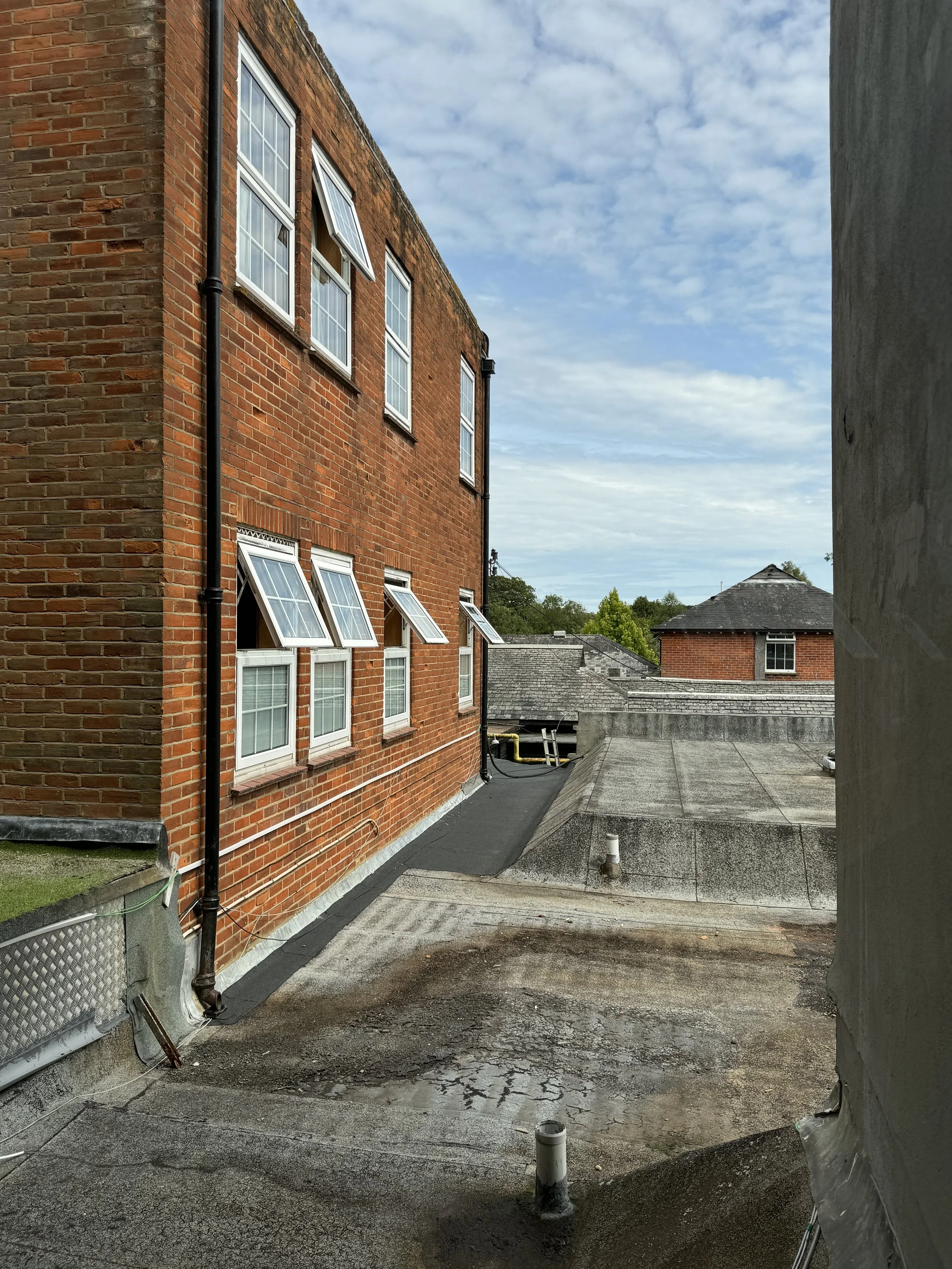 View of a flat rooftop with some vents, a black drainpipe along the side of a red brick building, and other rooftops in the background under a partly cloudy sky.