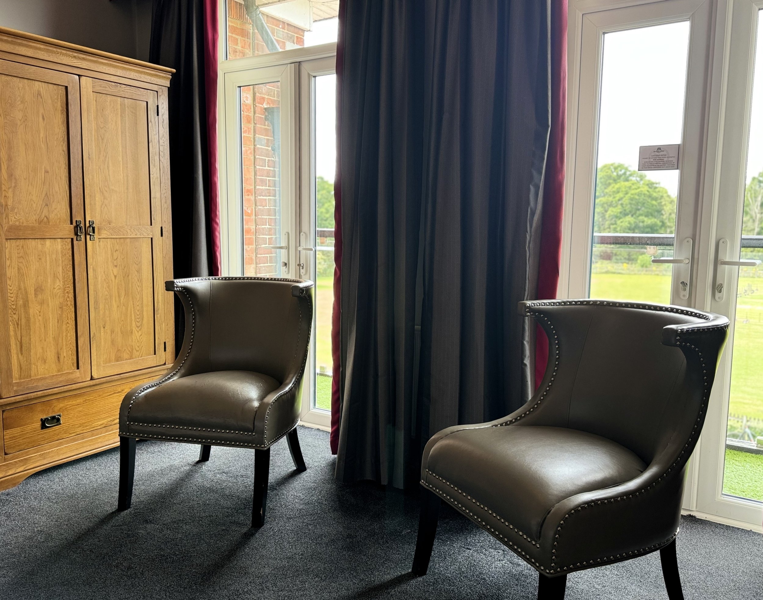 Interior of a room with two black leather armchairs, a wooden cabinet, and large windows with black and red curtains showing a green outdoor landscape.