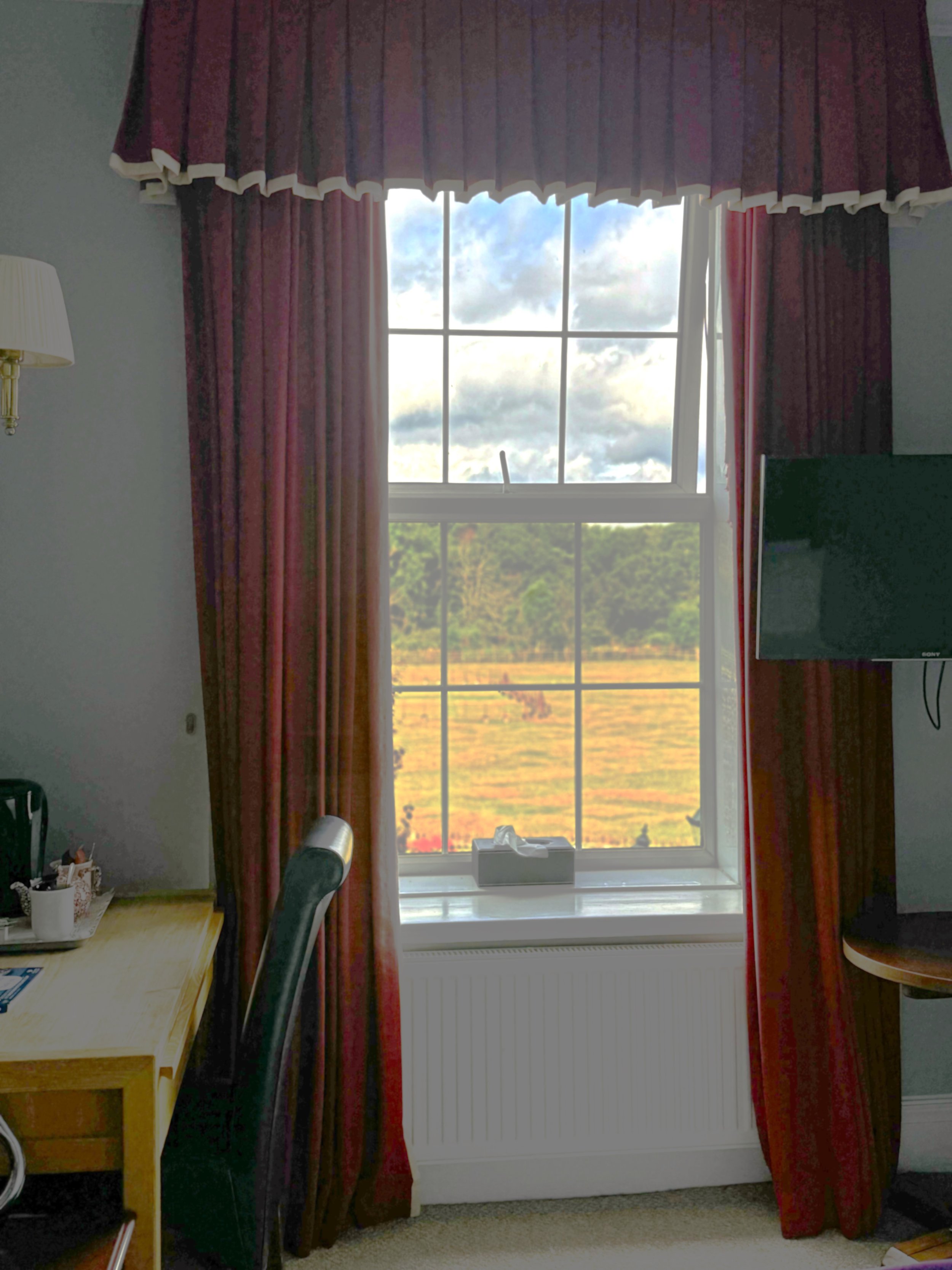View through a window with red curtains showing a scenic outdoor landscape with fields and trees and a cloudy sky.