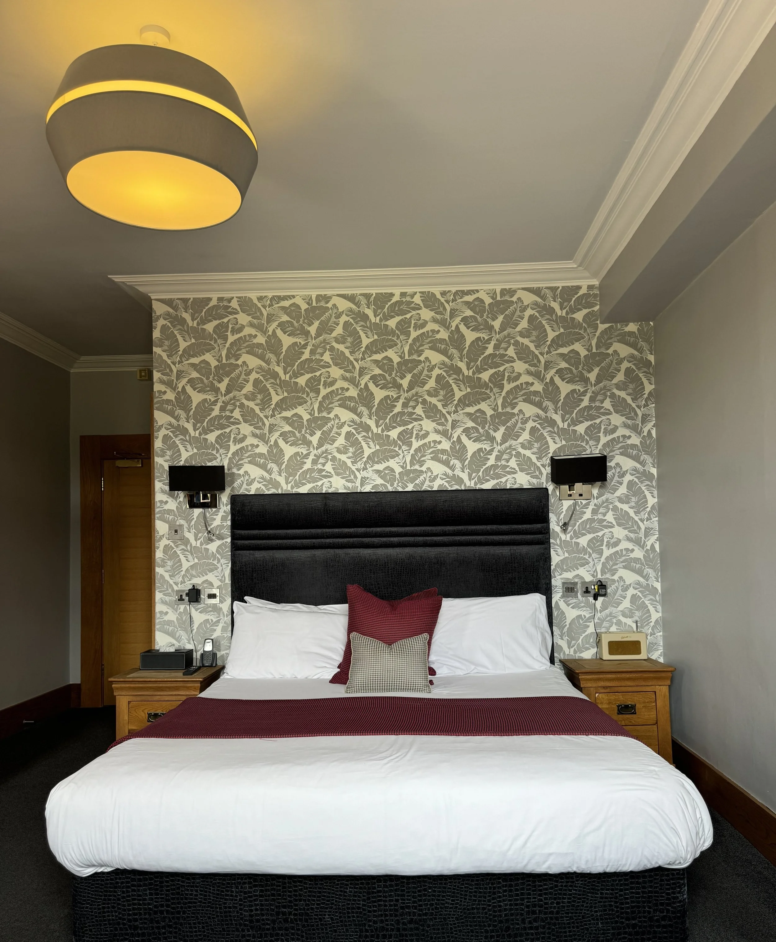 A hotel room featuring a bed with white sheets, a black headboard, and red and beige pillows. There are two black wall-mounted lamps, wooden side tables with telephones, and a patterned wallpaper with leaf motifs on the wall behind the bed. A ceiling