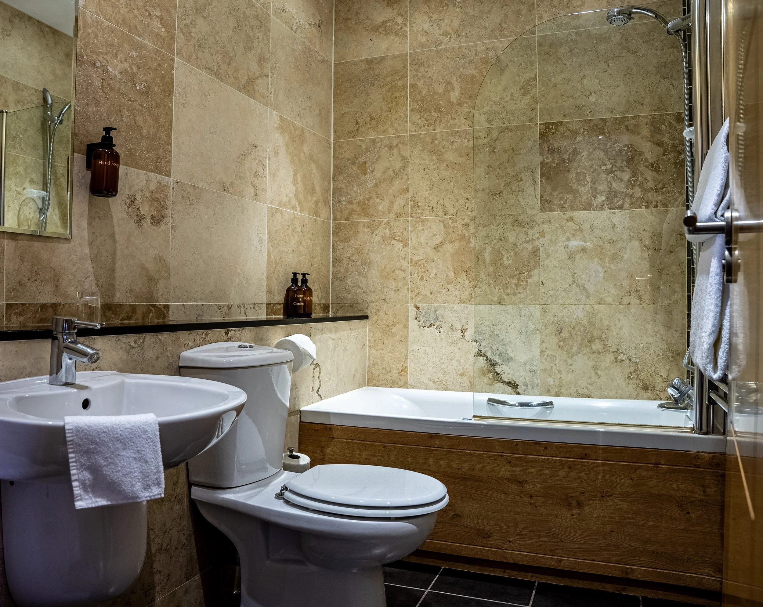 A bathroom featuring a sink with a white towel, a toilet, and a bathtub with a wooden panel, all with beige tiled walls.