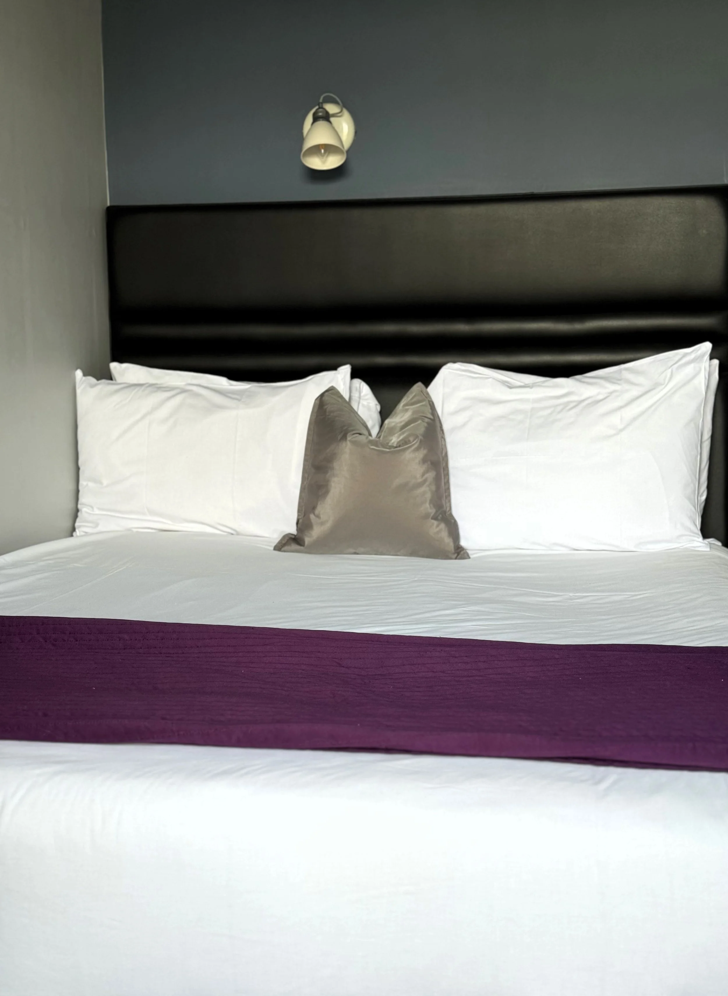 Hotel bed with white linens, a purple bed runner, and three pillows, one beige satin in front of two white pillows, with a dark headboard and a wall-mounted reading light above.