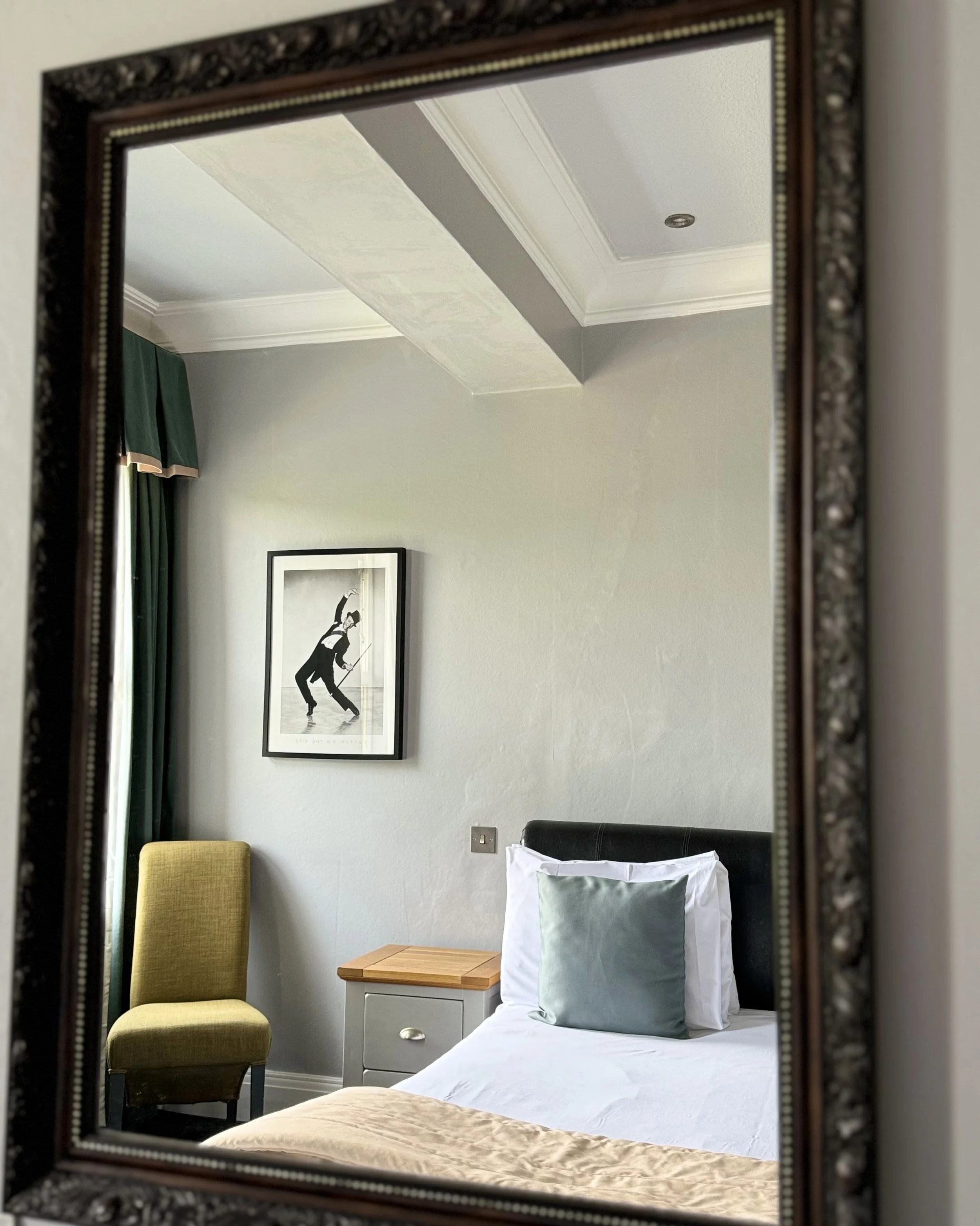 A hotel room is seen through a decorative mirror. The room has a bed with white sheets, a gray pillow, and a green pillow. To the left of the bed is a small nightstand with a wooden top, and a light green upholstered chair is placed next to the night