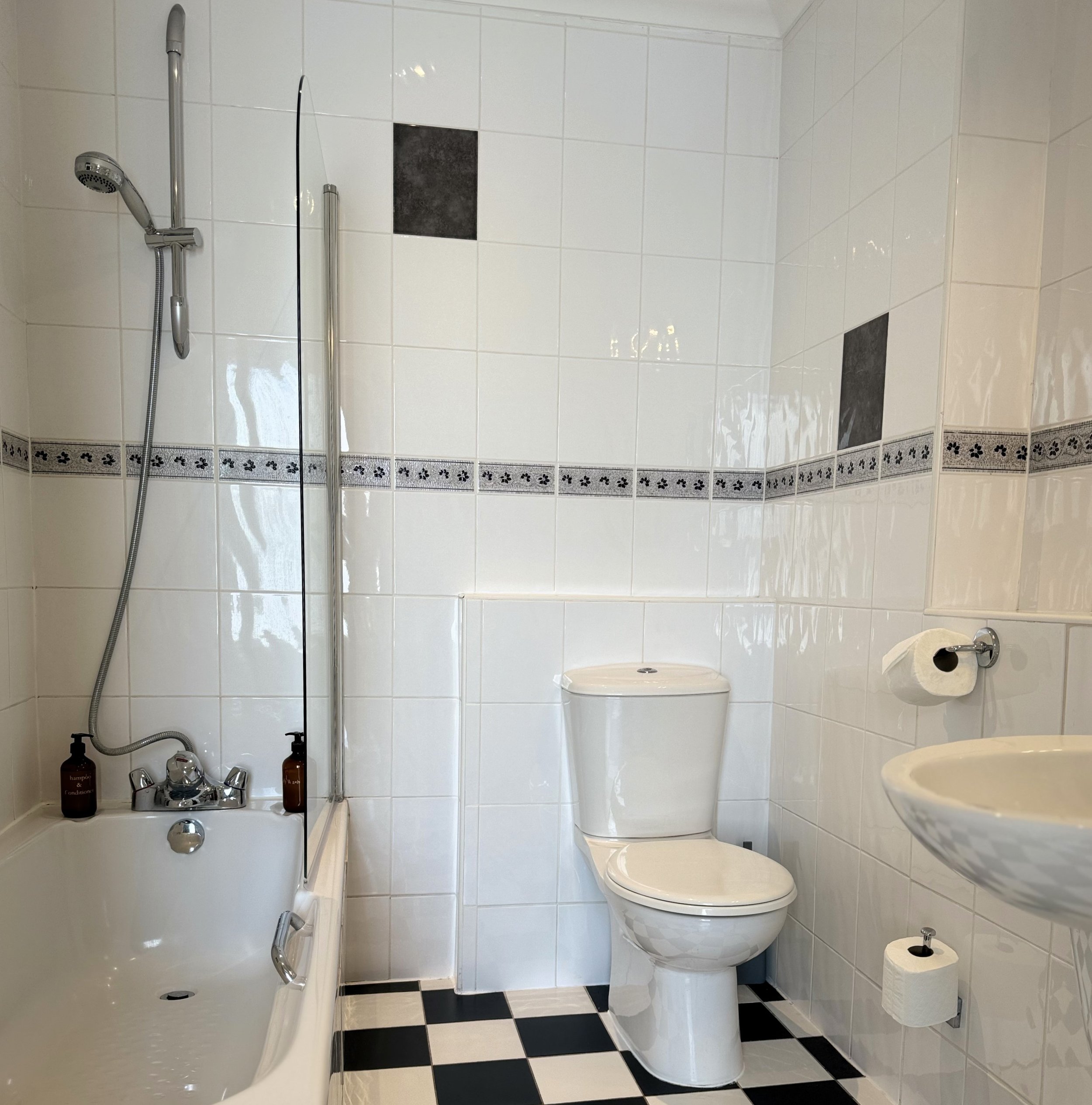 A small bathroom with white tiled walls, black and white checkered floor, and a white toilet. There is a bathtub with a handheld showerhead and two bottles of soap or shampoo on the edge. A partial view of a small wall-mounted sink with toilet paper 