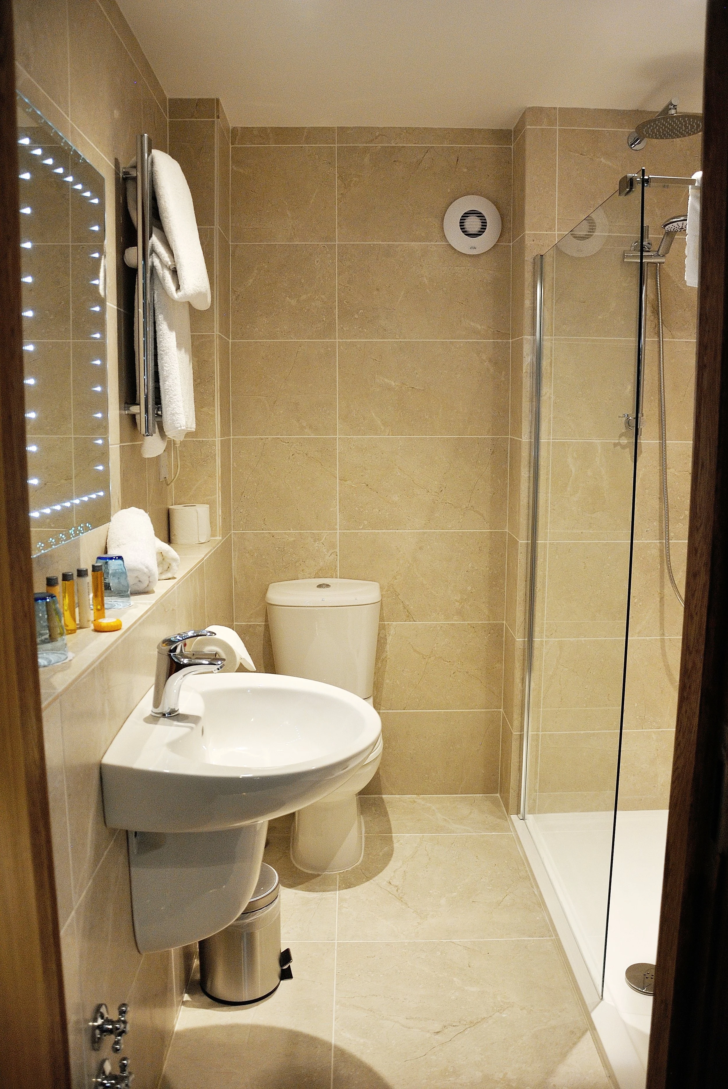 A small bathroom with beige tiled walls and floor, a white sink, a toilet, a walk-in shower with a glass door, towels on a heated towel rack, toiletries on a shelf, and a mirror with built-in lighting.