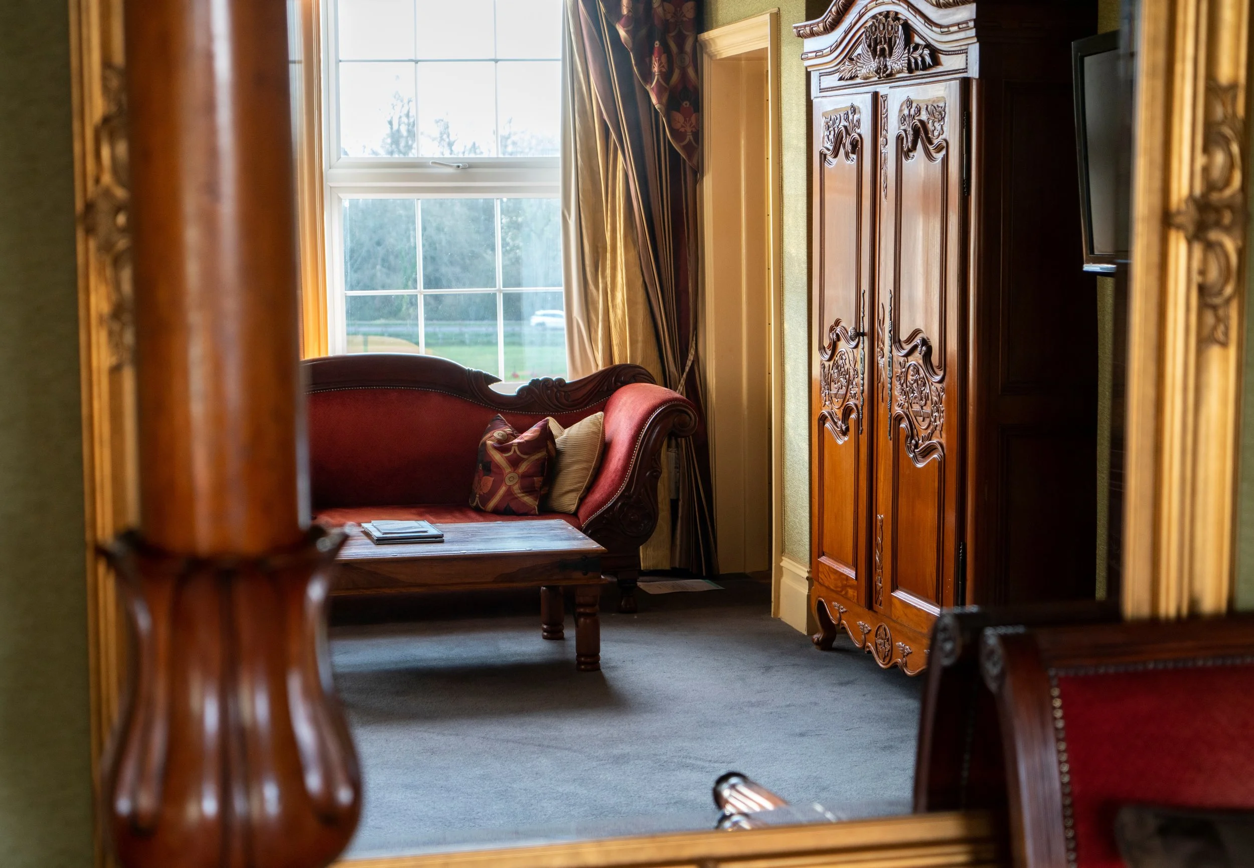 A cozy room with antique furniture, including a carved wooden armoire, a red velvet sofa with pillows, and a wooden coffee table with magazines. There's a large window with curtains, letting in natural light, and a wall-mounted TV is visible on the r