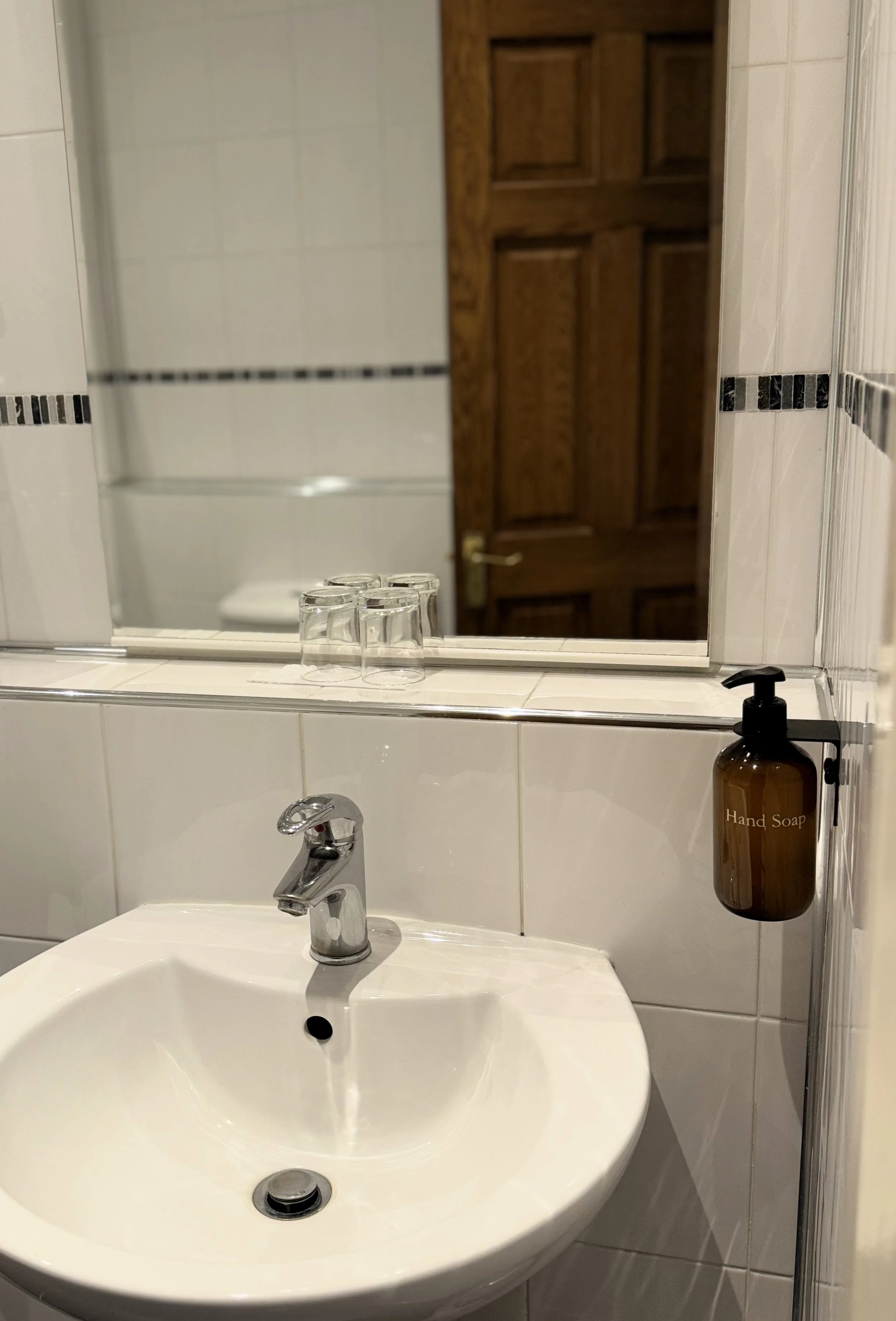 Bathroom sink with a mirror above it, a hand soap dispenser on the wall, and three glasses on the mirror's ledge, with a wooden door reflected in the mirror.