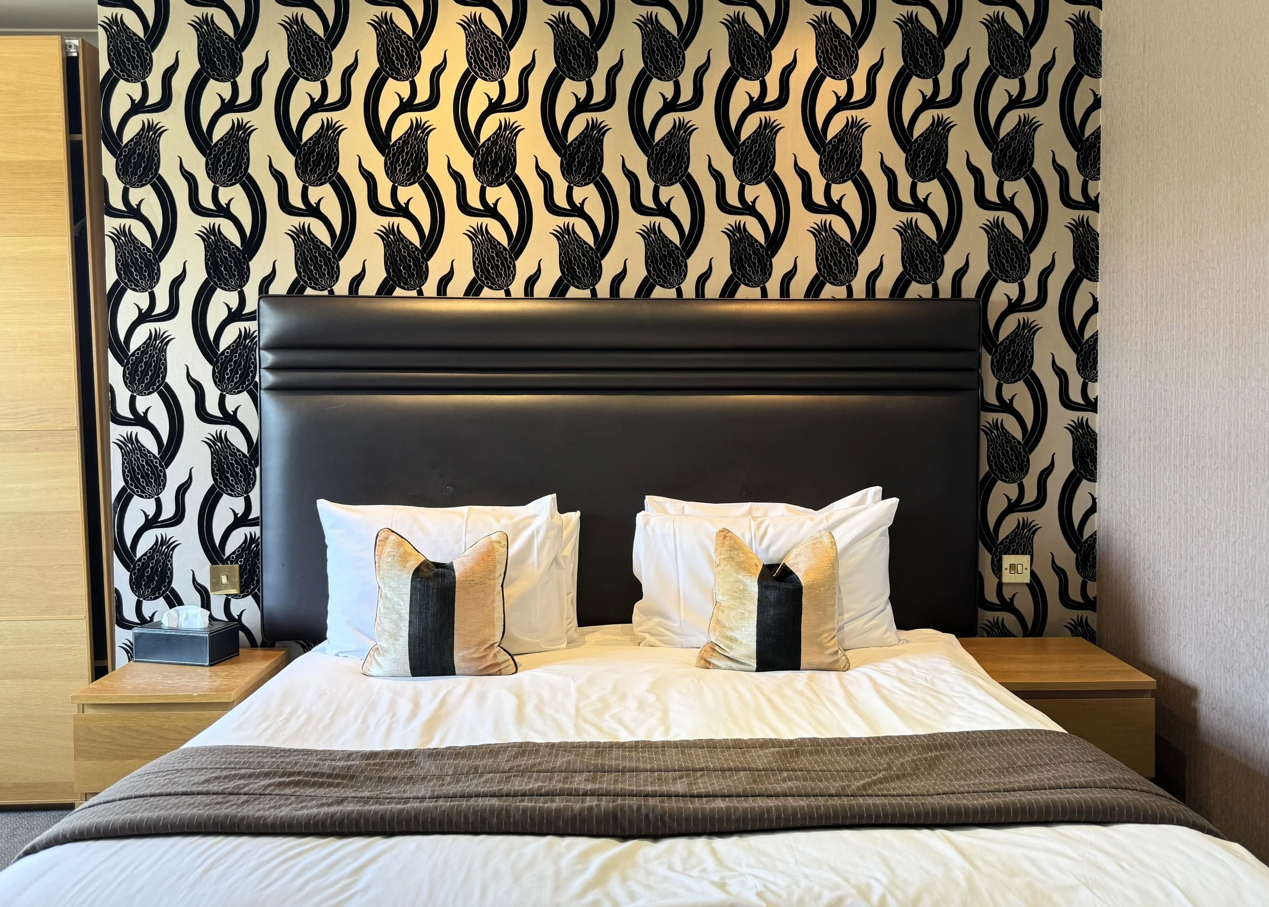 A neatly made bed with white sheets, two white pillows with decorative covers, and a dark brown bed runner. The bed has a black padded headboard, flanked by wooden nightstands, with a box of tissues on the left and an electrical outlet on the right. 