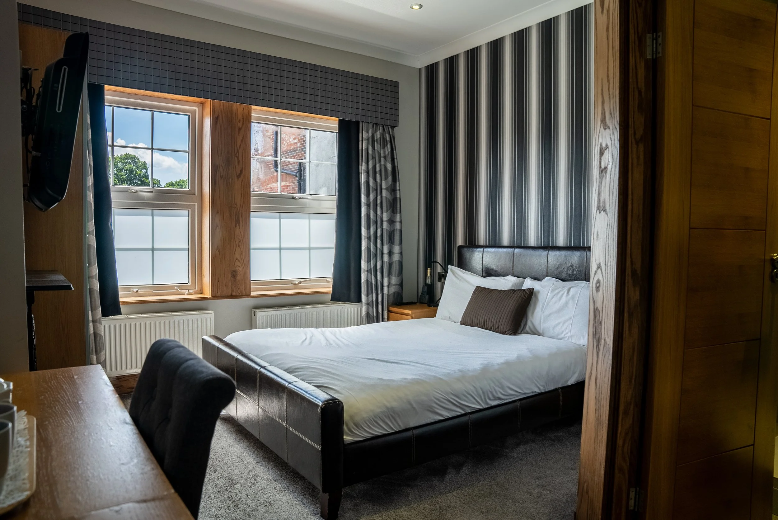 A cozy hotel bedroom with a large bed, dark leather headboard, white bedding, brown pillow, and a window with patterned curtains, wooden trim, and a view of a brick building outside.