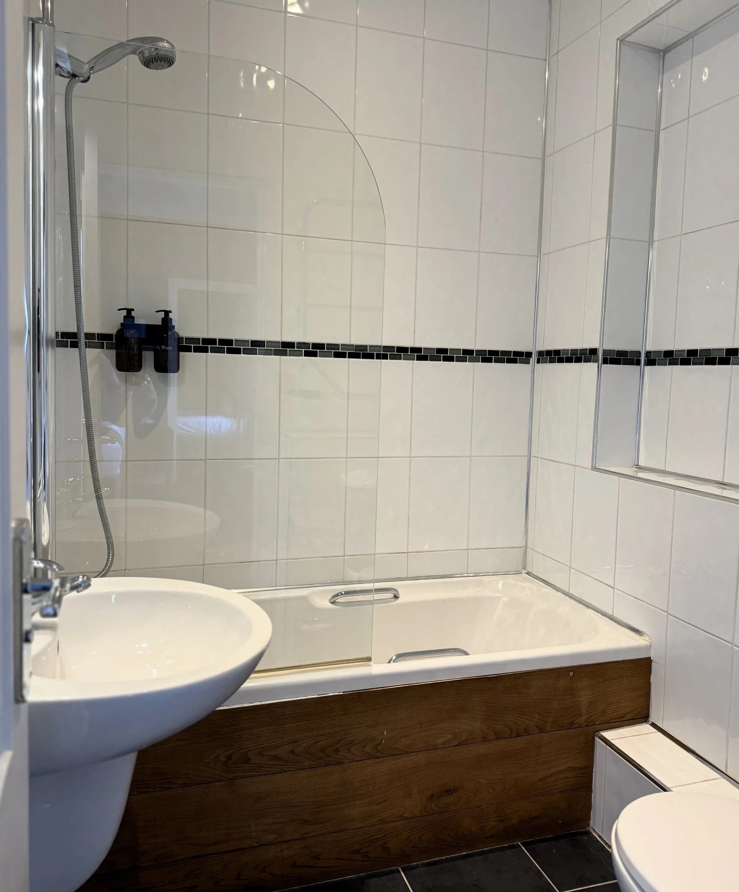 Bathroom with a bathtub, showerhead, and a small sink, with white tiled walls and a glass shower screen.