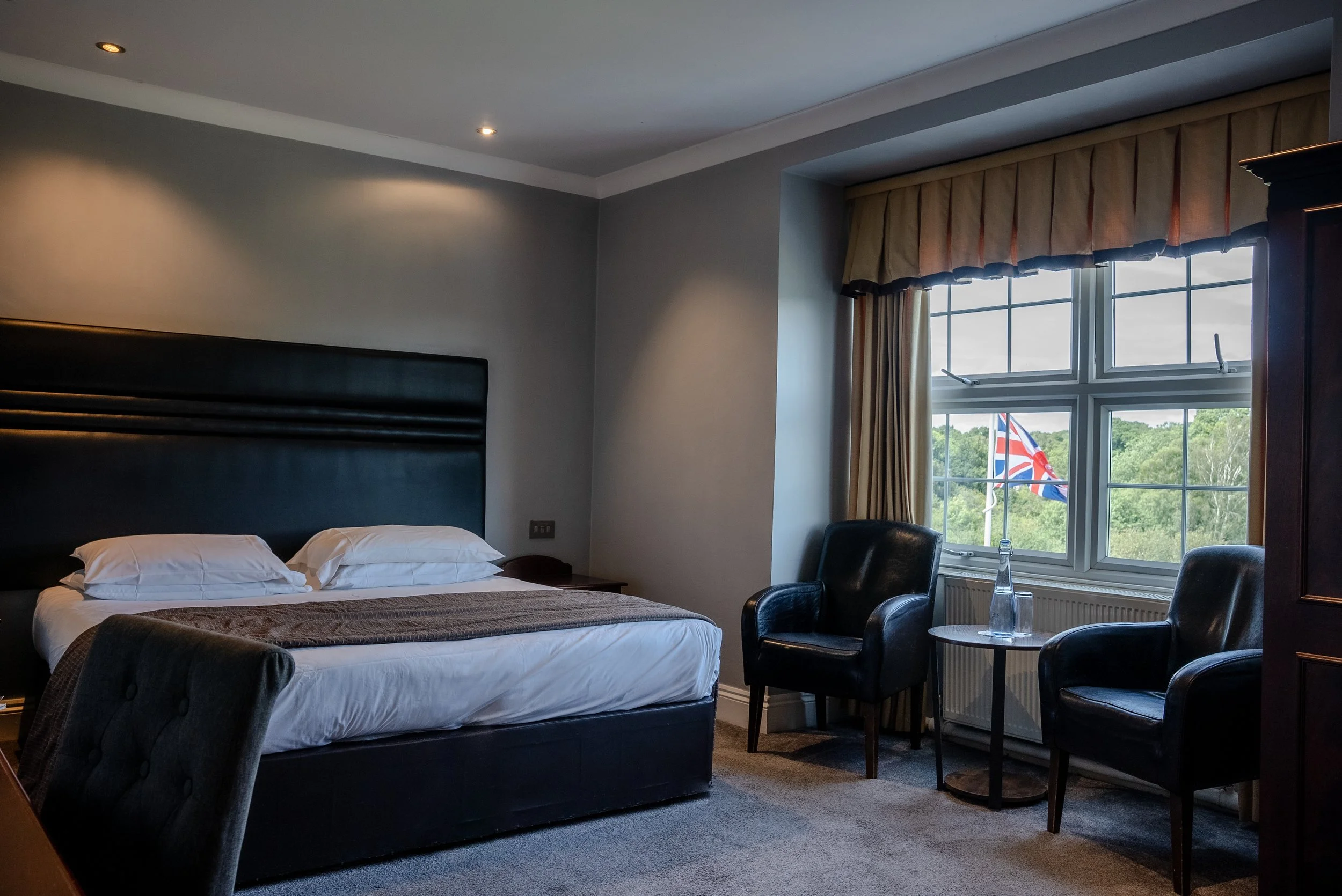 Hotel room with a large bed, two black armchairs, a side table with a bottle of water, and a window with cream-colored curtains and a Union Jack flag outside.