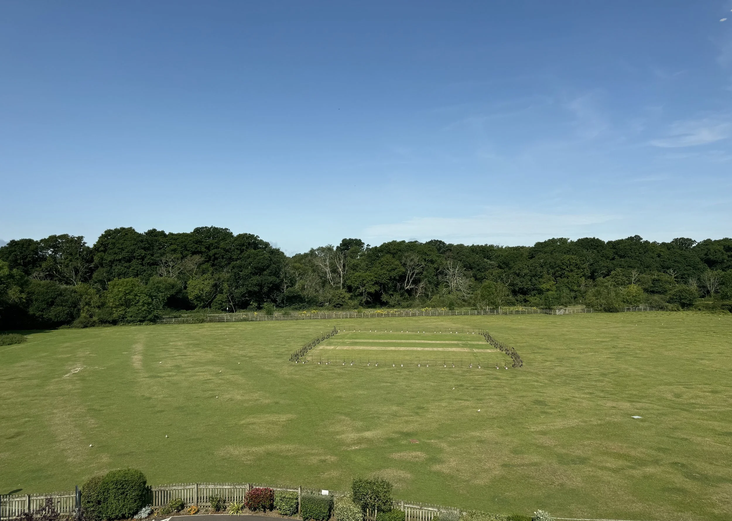 Wide grassy field with a lined target shooting range in the center, surrounded by trees and a fence, under a clear blue sky.