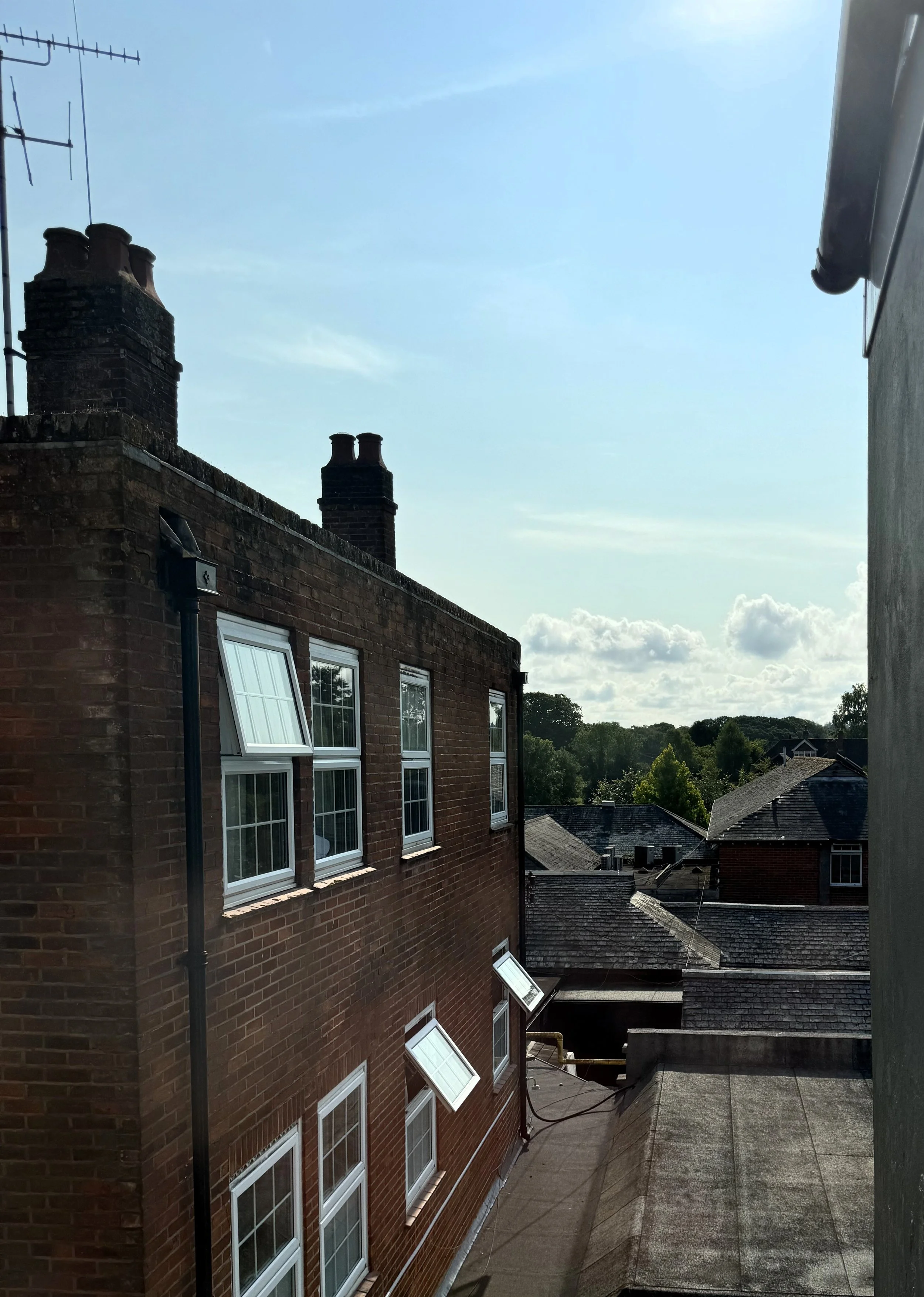 View from a rooftop showing a brick building with multiple windows and open window panes, rooftops of neighboring buildings, trees in the background, and a partly cloudy sky with sunlight.