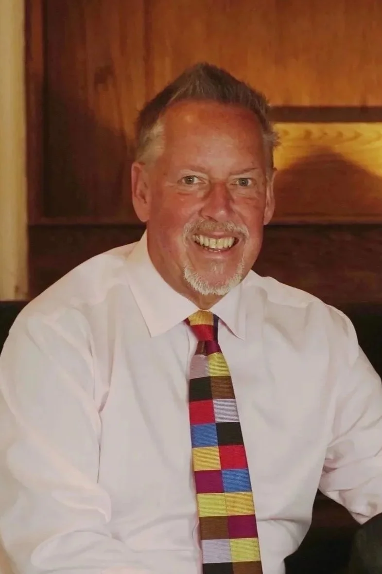 A middle-aged man with short gray hair, a goatee, and a broad smile, wearing a floral shirt.