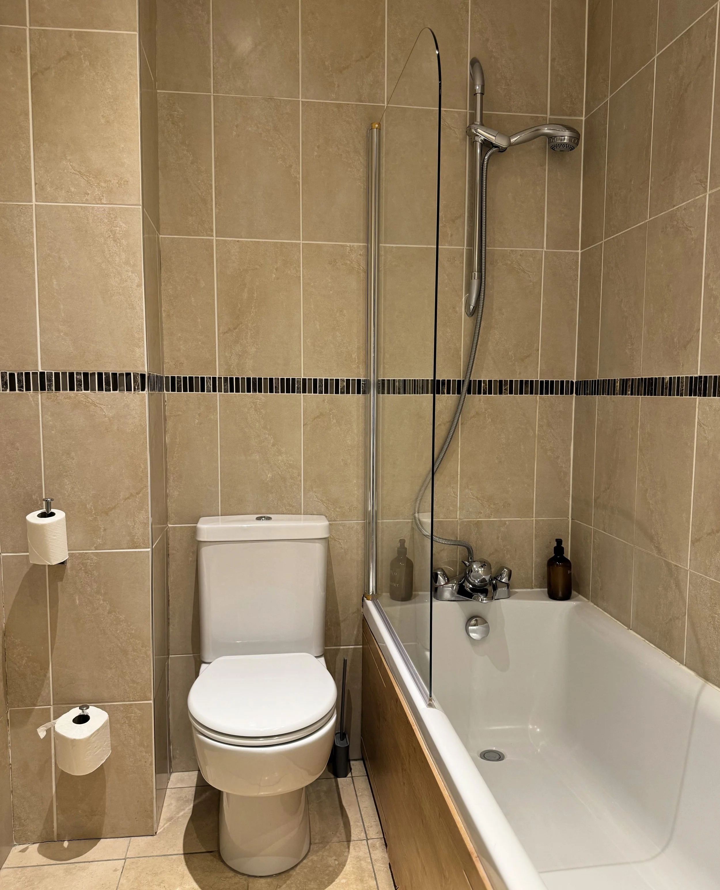 Bathroom with beige tiled walls, a toilet, and a bathtub with a shower head and glass partition.