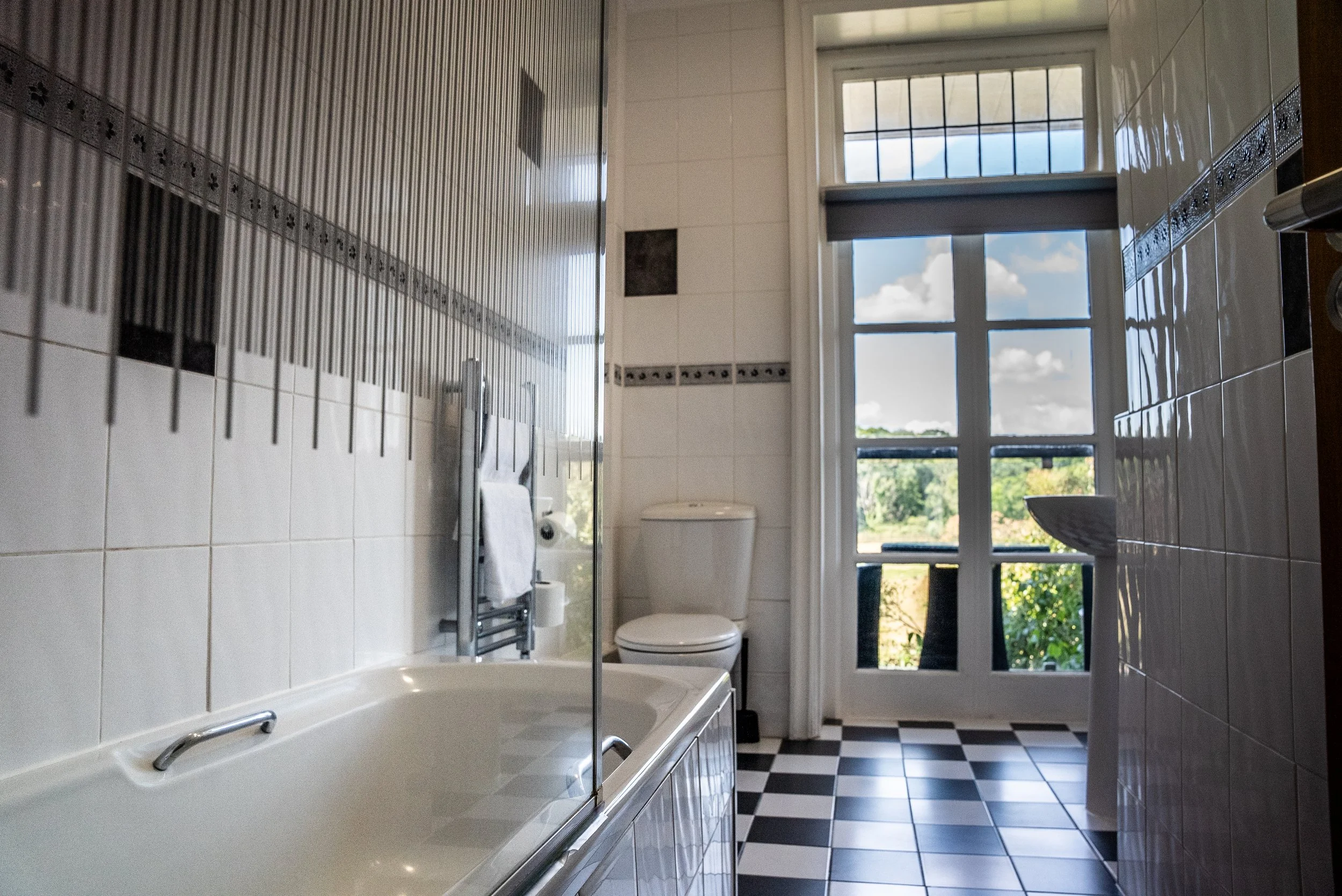 Bathroom with bathtub, toilet, sink, large window showing outdoor scenery, checkered black and white floor tiles, white tiled walls with decorative border.