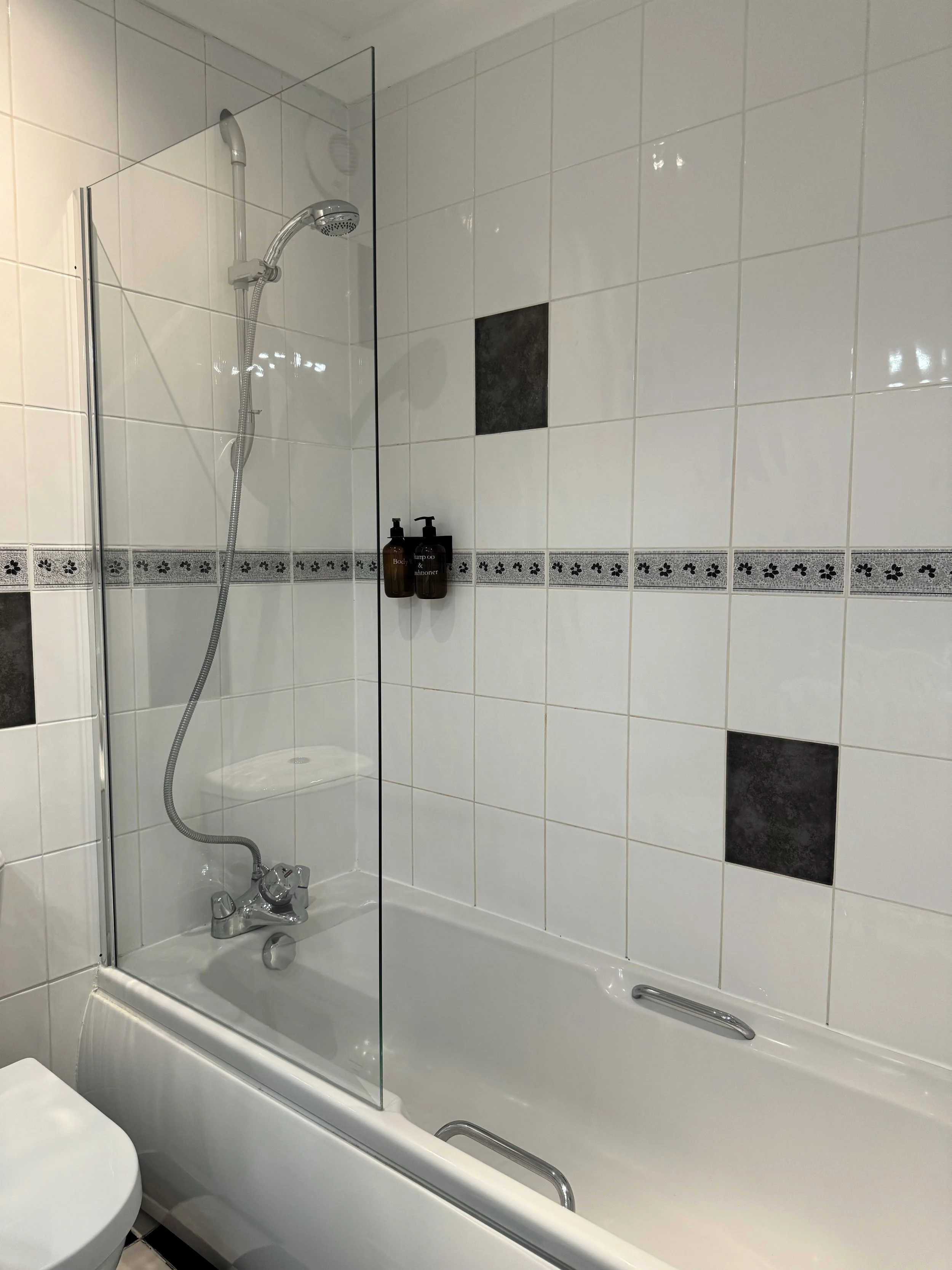 Bathroom with shower and bathtub, white tiled walls with black decorative tiles, shower with handheld sprayer, glass shower screen, wall-mounted soap or shampoo dispensers, black tile accent in wall