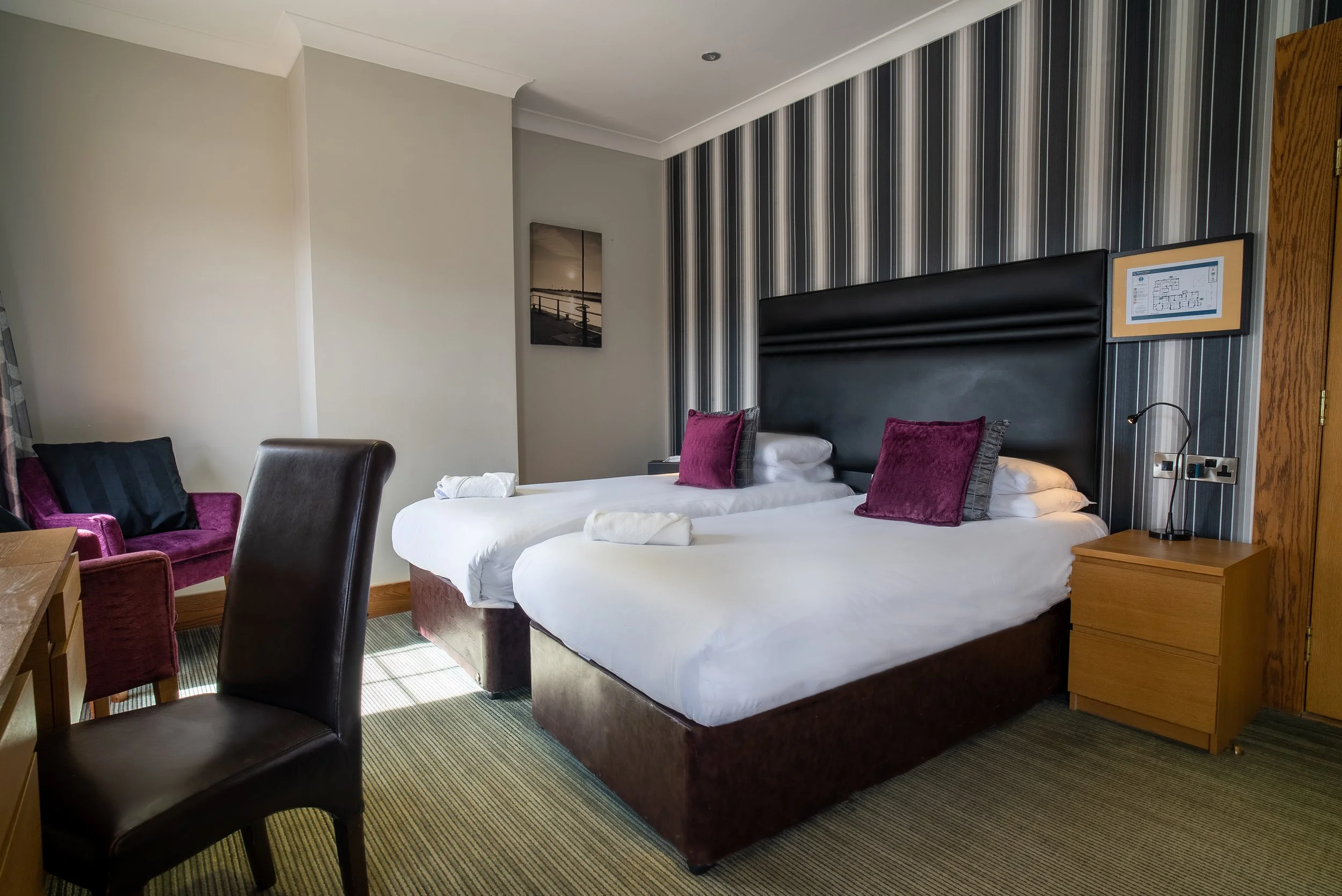 Hotel room with two single beds, each with a burgundy pillow, a bedside table with a reading lamp, a purple armchair, a black leather chair, and a wooden desk. The room features a striped wallpaper behind the beds and a framed picture on the wall.