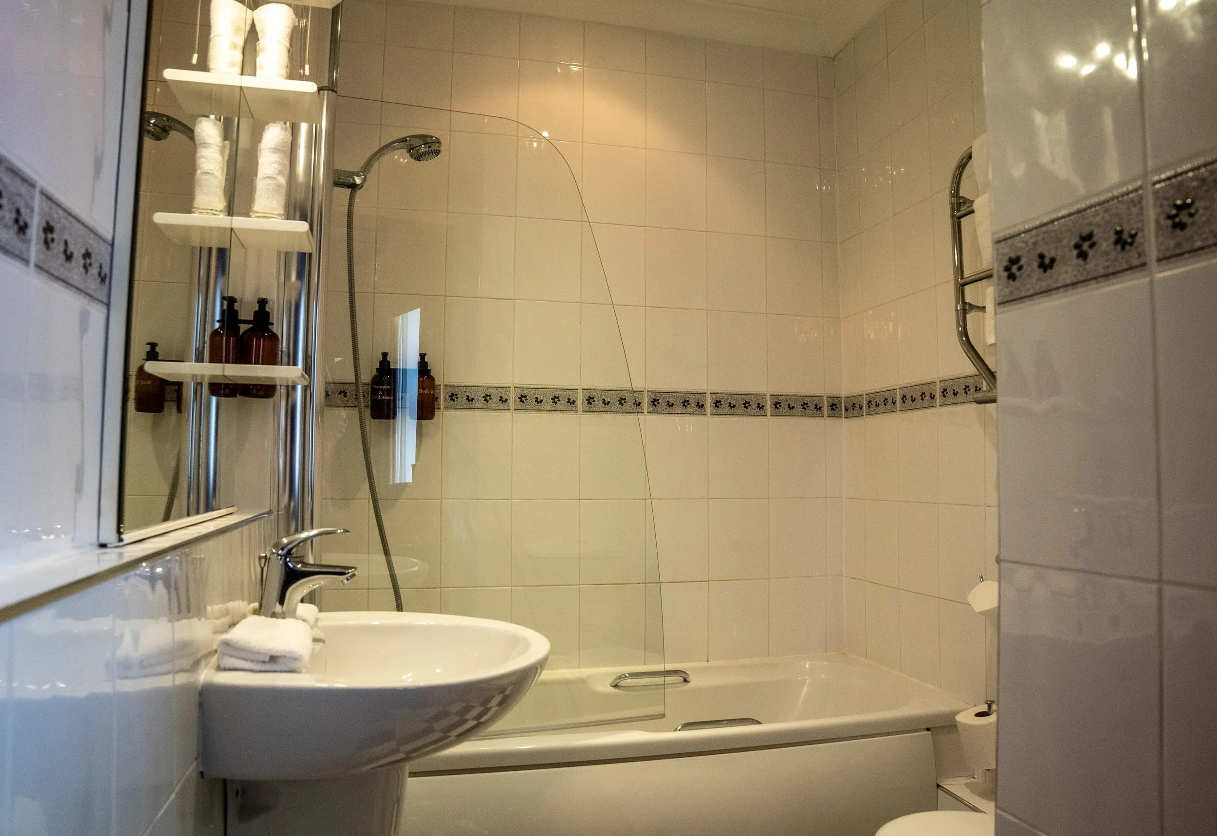 A small bathroom with beige tiled walls, a bathtub with a glass shower screen, a corner shelf with toiletries, a small white sink with a towel, and a toilet paper holder with a roll of toilet paper.