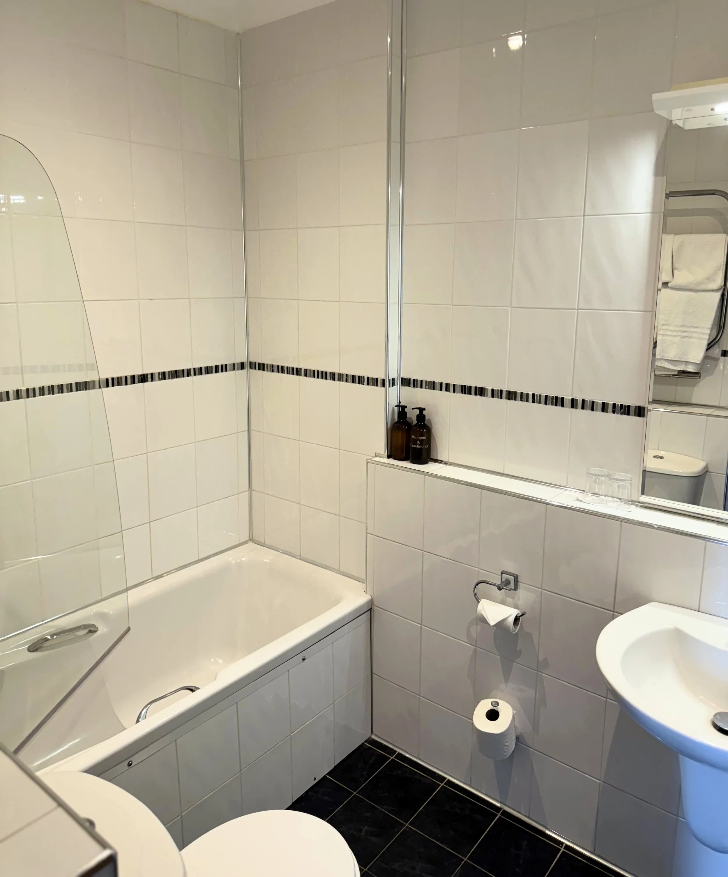 Bathroom with bathtub, toilet, sink, mirror, and wall tiles, with toiletries on a ledge and rolled towels on a rack.