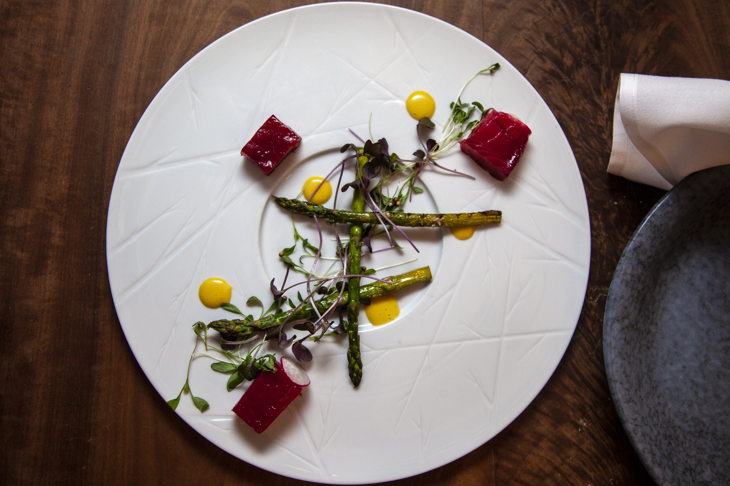 A plate with asparagus, microgreens, red jelly cubes, and yellow sauce drops on a wooden table.