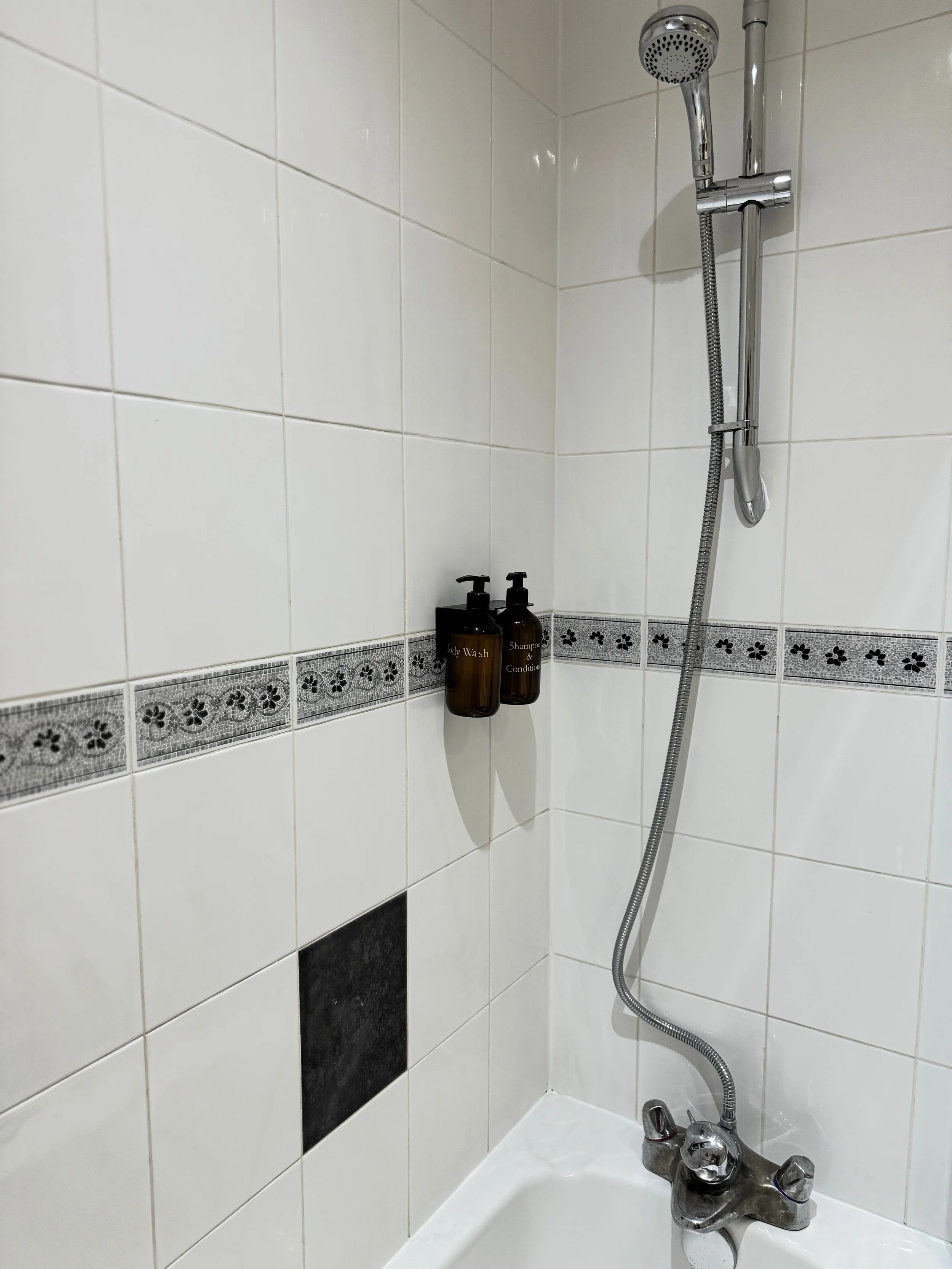 Shower area with white tiled walls, a black tile accent, a wall-mounted shower head, and two brown bottles labeled 'Body Wash' and 'Shampoo & Conditioner'