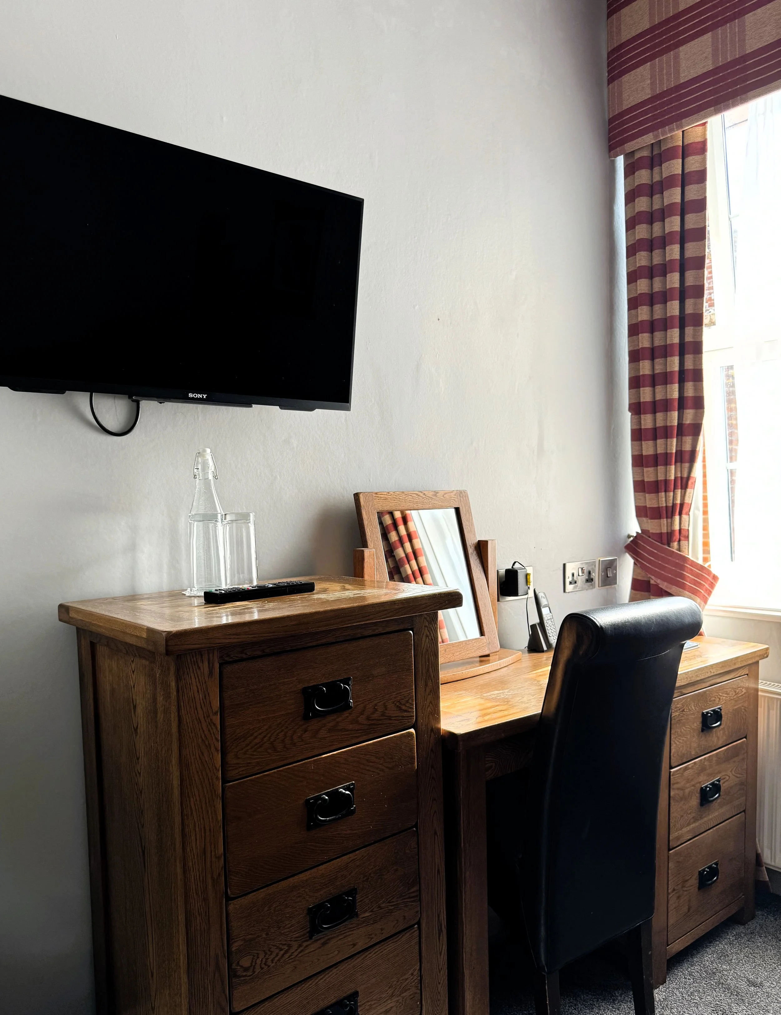 A hotel room workspace with a wooden desk, a black chair, a mirror, a TV mounted on the wall, and a window with red and beige striped curtains.