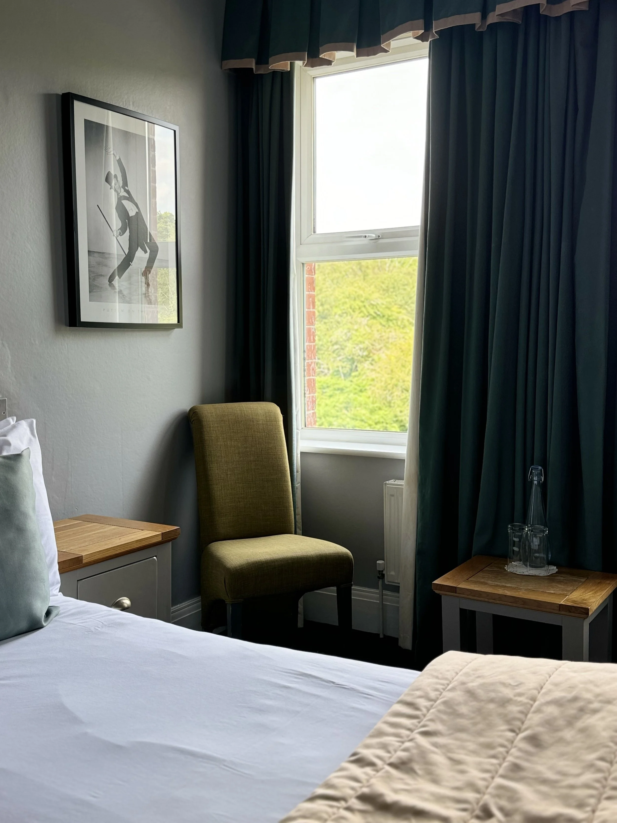 Room with a bed, bedside table, green chair, window with green trees outside, and wall art of a person playing golf.
