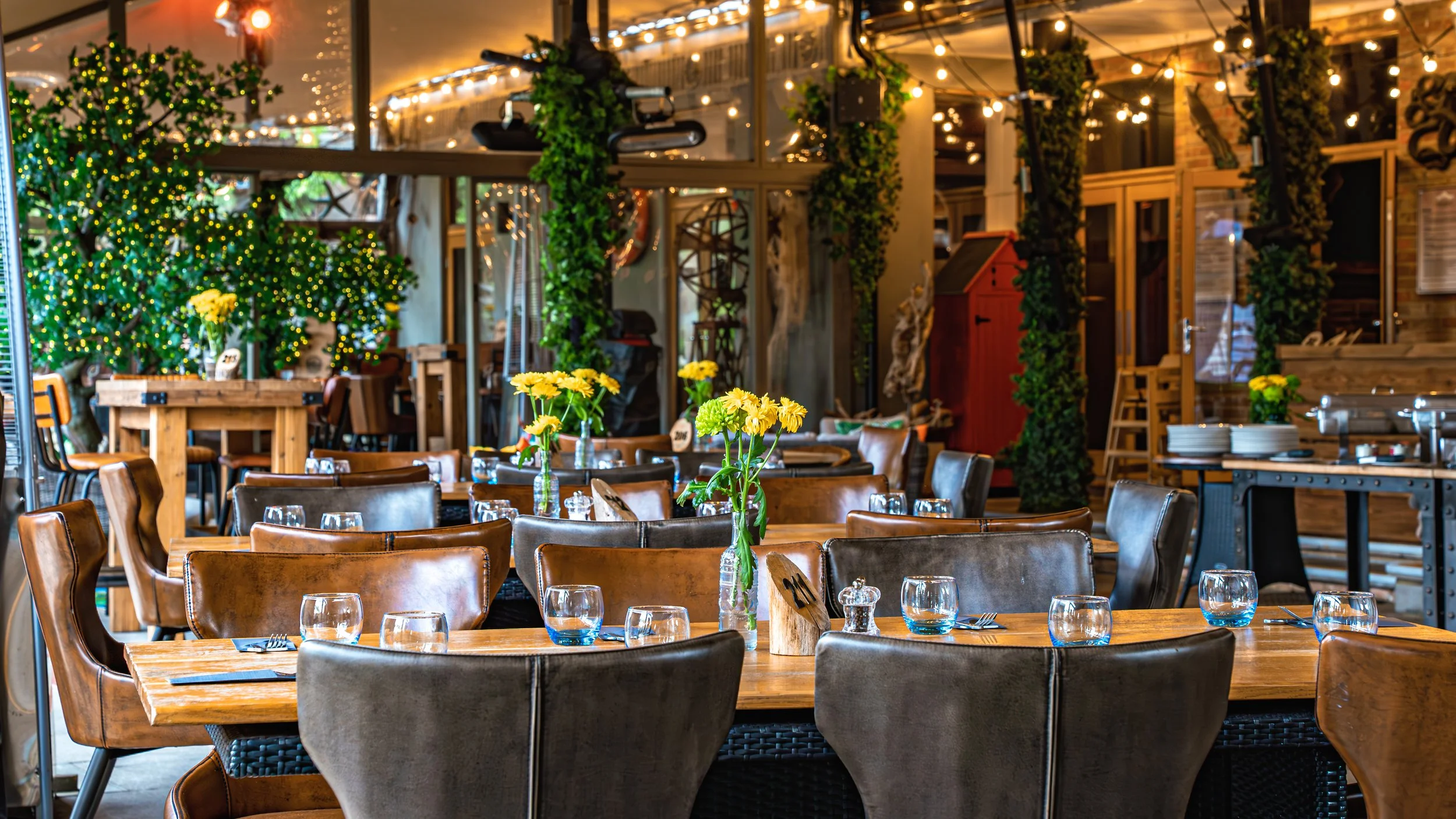 A long dining table set for a meeting or dinner with multiple flower arrangements in vases and place settings with notebooks and pens. A chandelier hangs from the ceiling, and a television is visible in the background.