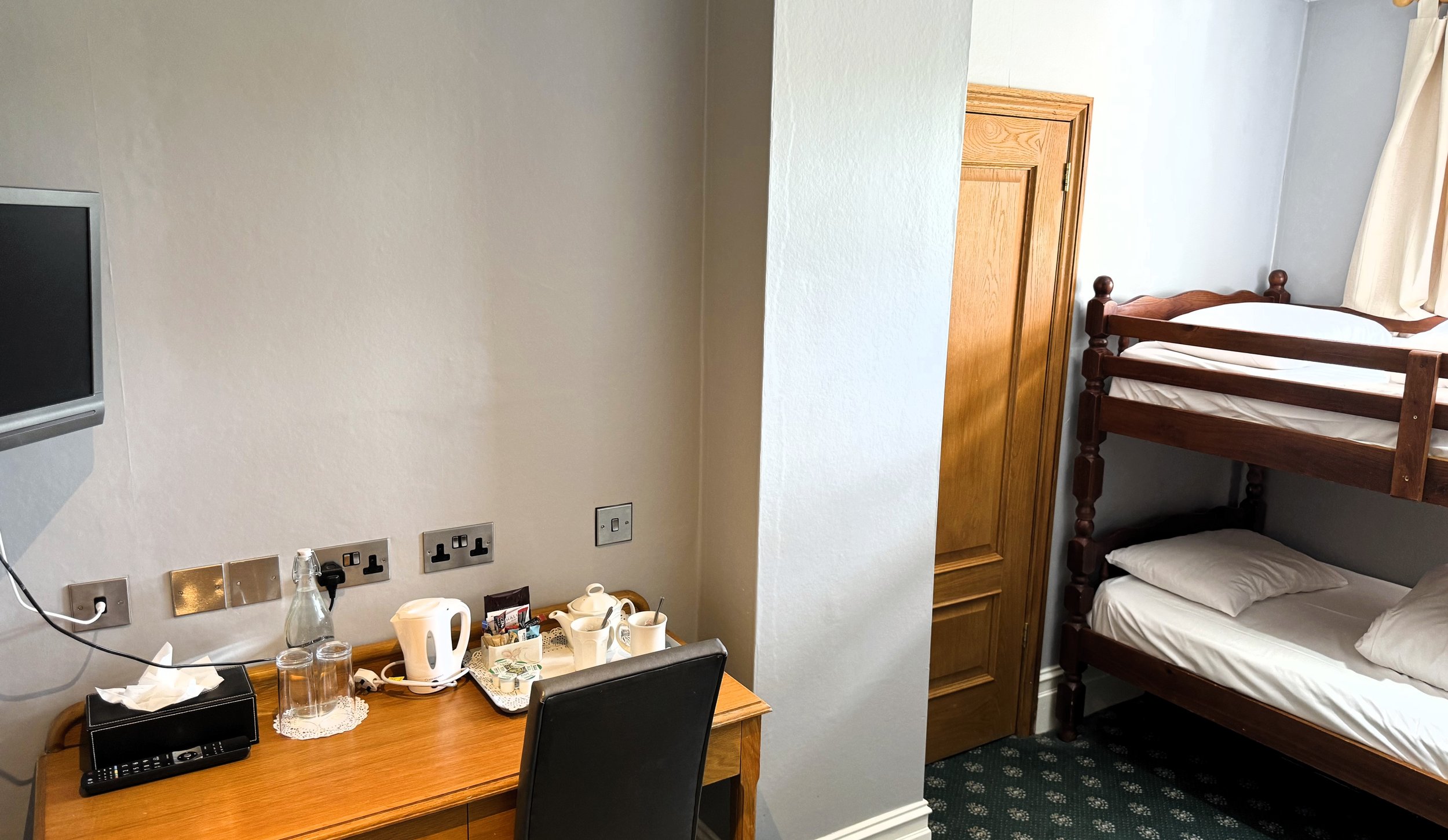 Part of a hotel room showing a wooden desk with a television remote, tissue box, glasses, teapot, cups, and packet of tea or coffee; a black chair; a wooden bedroom door; a bunk bed with white bedding and pillows; a window with white curtains; over c