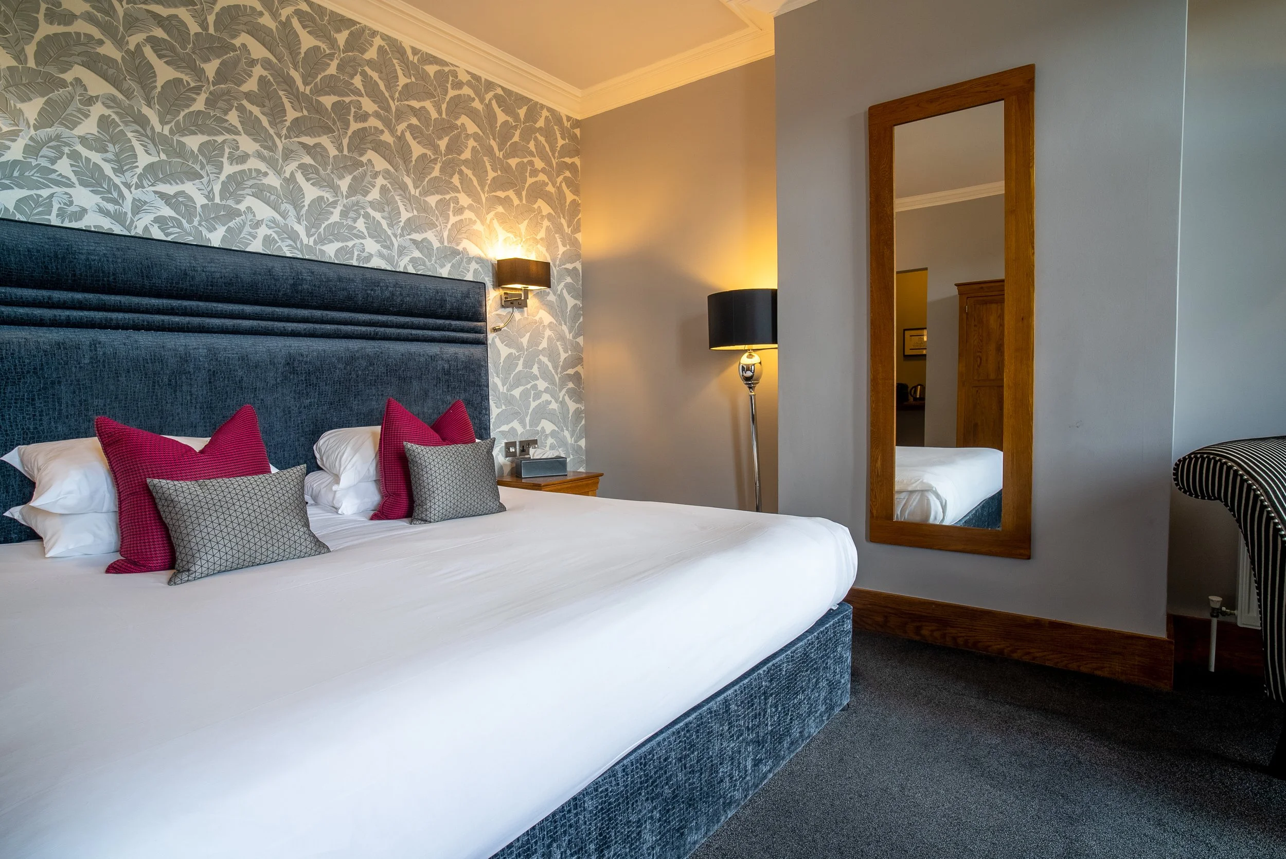 A neatly made bed with a blue upholstered headboard, white bedding, and decorative red and grey pillows. A wall-mounted lamp and a floor lamp are beside the bed. There is a wooden-framed mirror on the light grey wall and a wooden nightstand with a sm