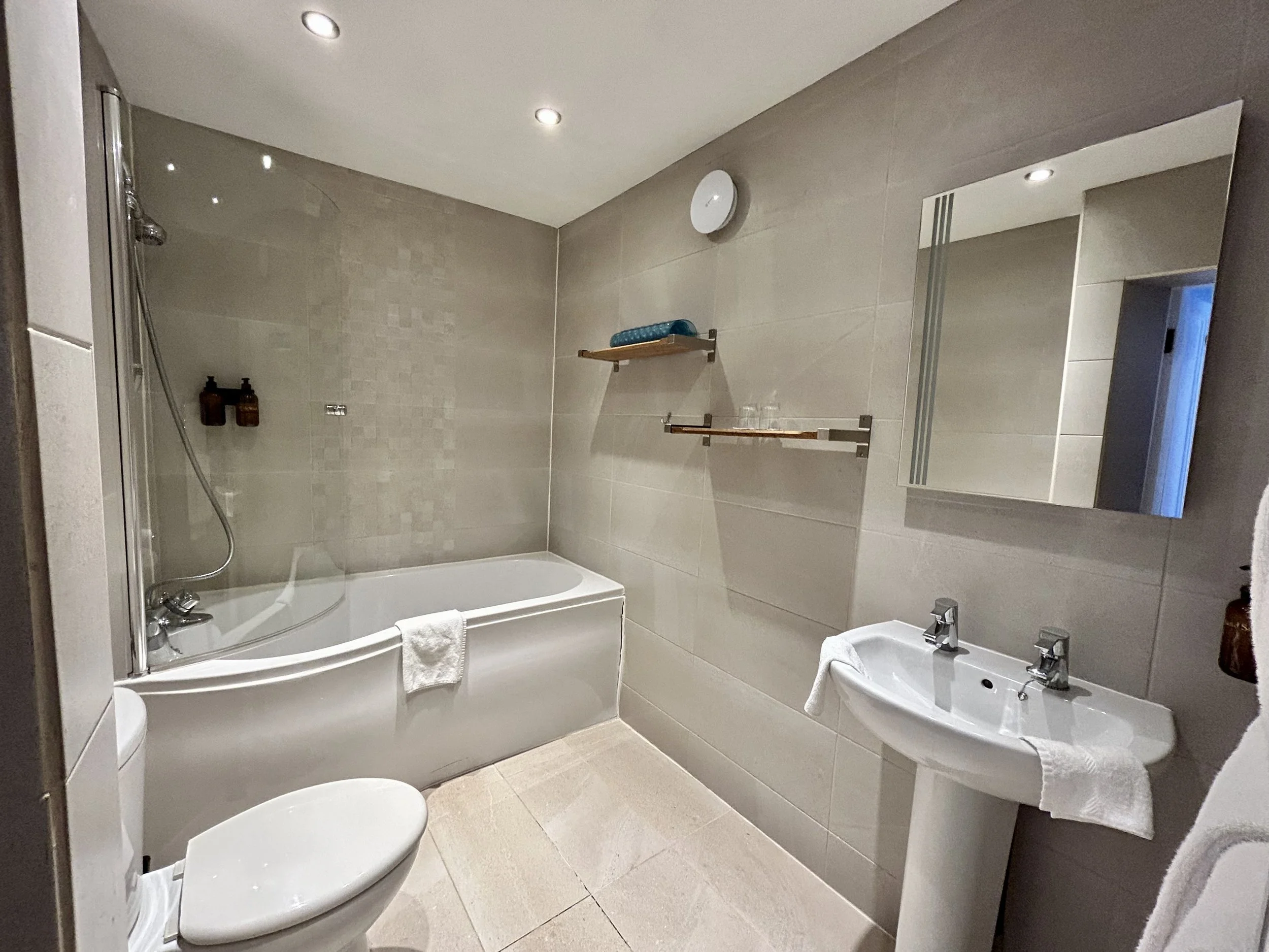 Modern bathroom with a bathtub, toilet, and sink, beige tiled walls, a mirror, and shelf with glasses.
