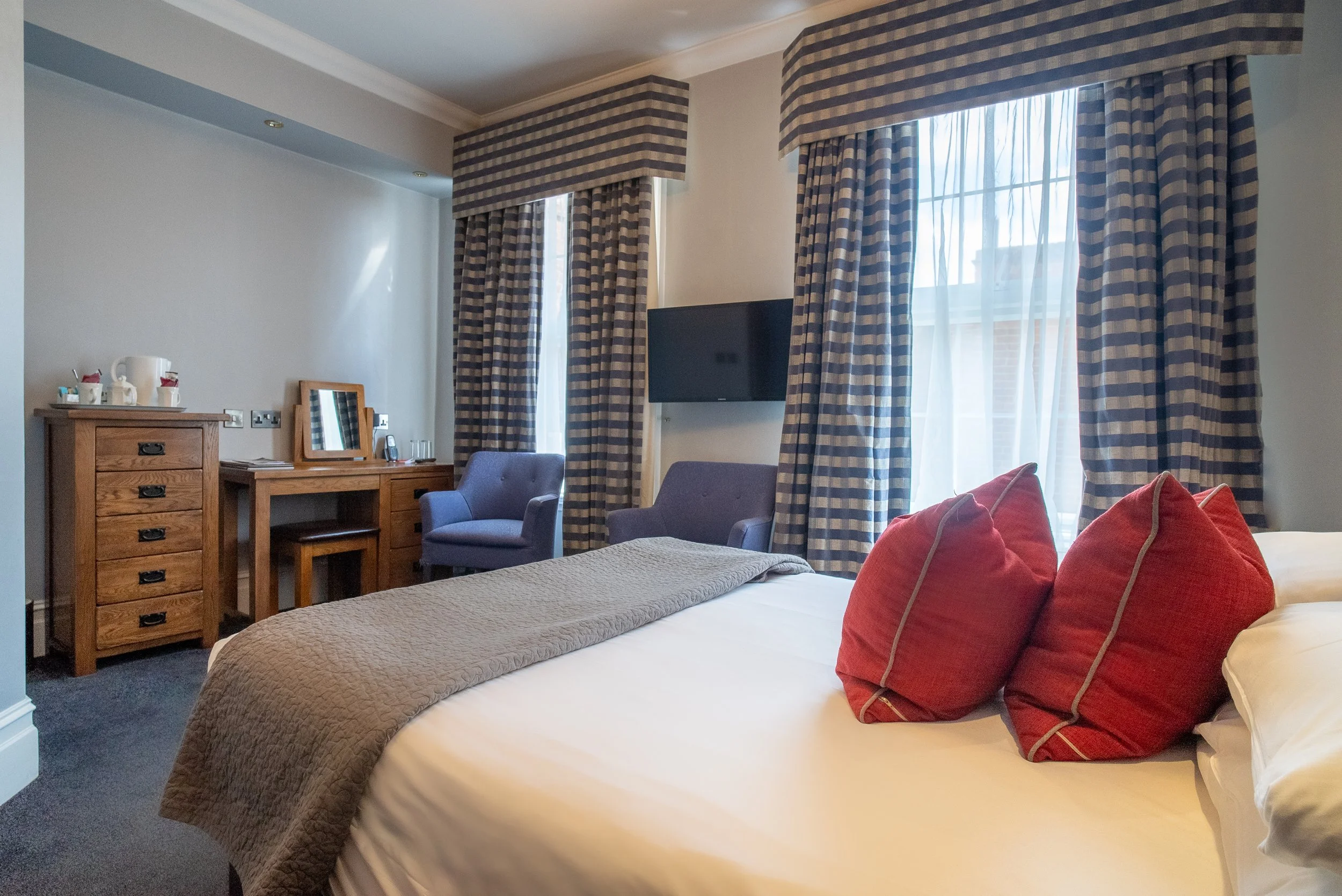 A cozy hotel room with a bed featuring red pillows, a beige bedspread, and large windows with plaid curtains. There is a wooden dresser with tea set and cups, two blue armchairs, and a mounted flat-screen TV.