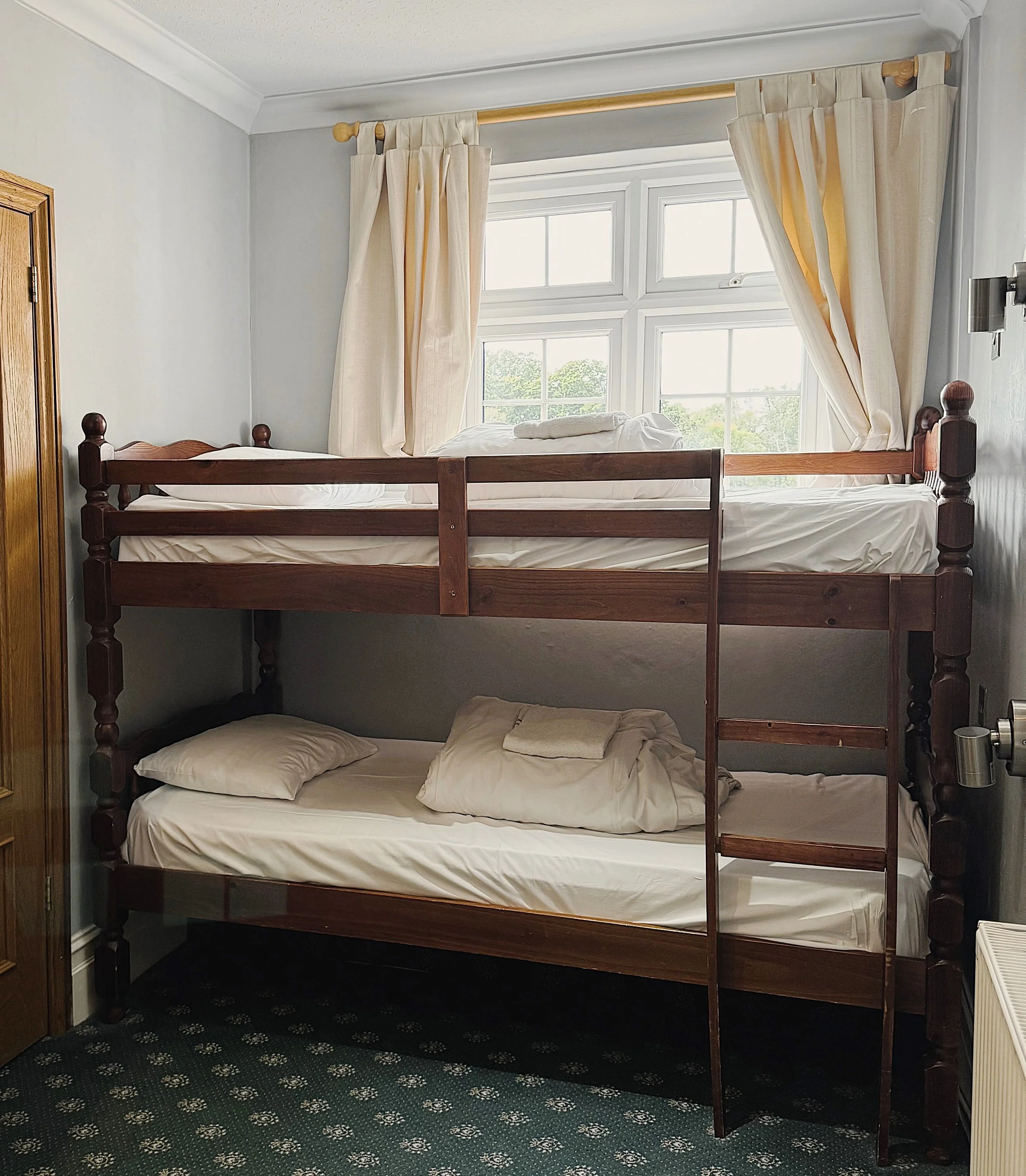 A room with a wooden bunk bed positioned in front of a window with cream-colored curtains, and white bedding on both beds.