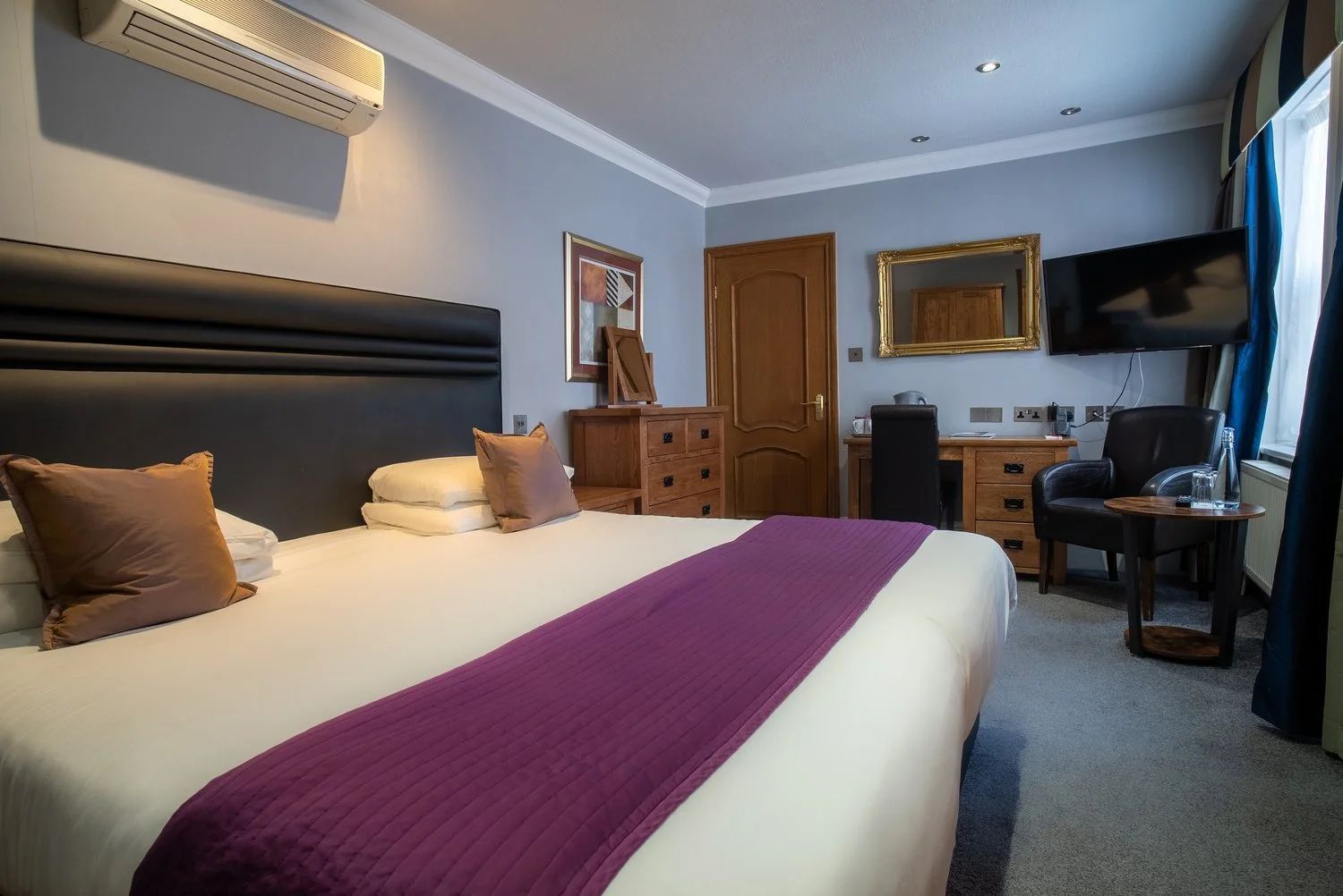 Hotel room with a large bed, brown pillows, purple bed runner, wooden furniture, a flat-screen TV, armchair, window with curtains, and an air conditioning unit.