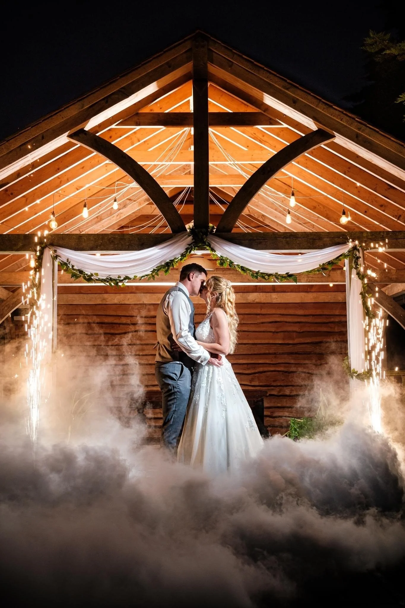 Couple in wedding attire dancing in a decorated outdoor wooden pavilion at night with sparklers and fog effect.