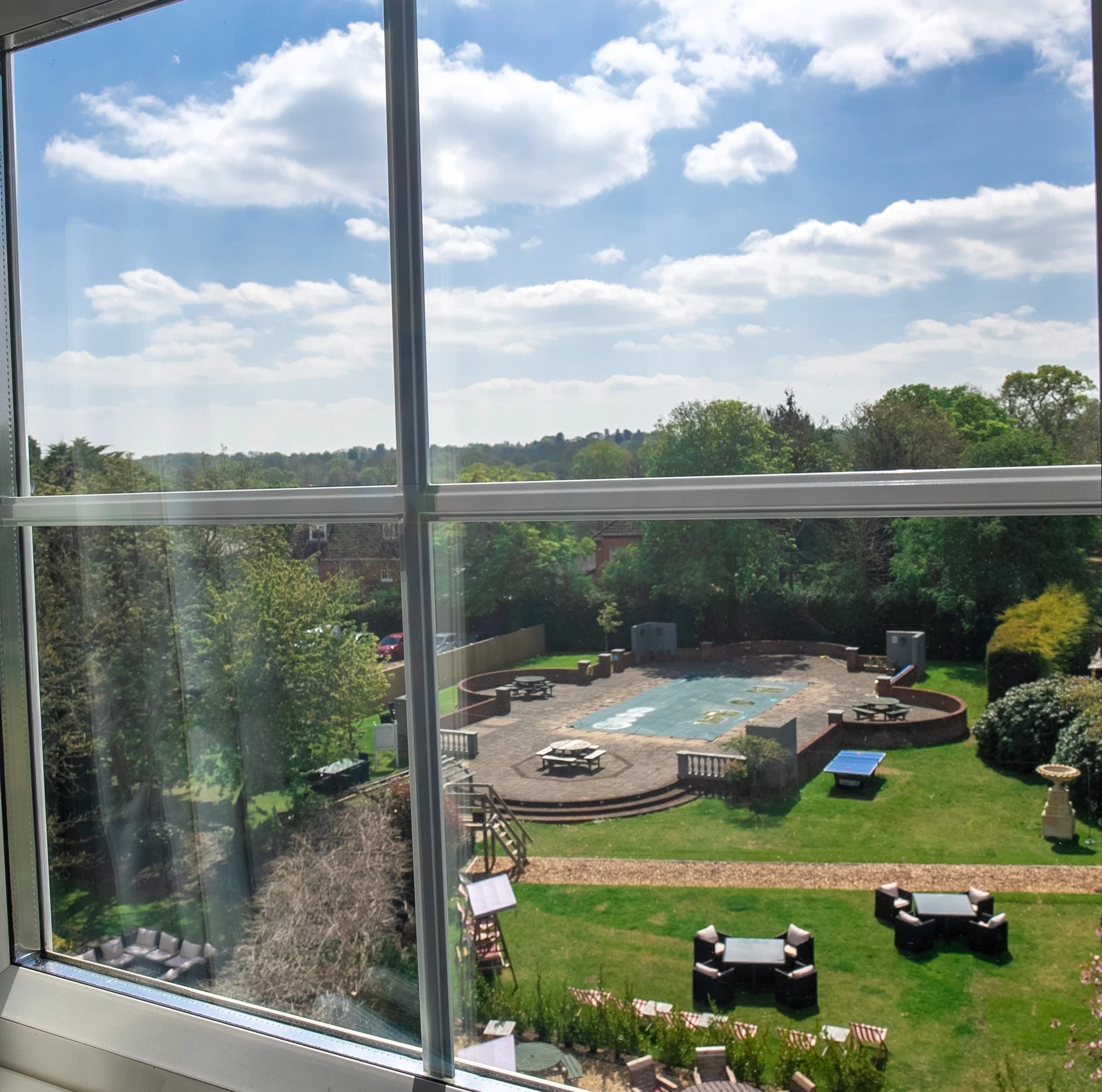 View of a backyard with a large patio area, round tables with umbrellas, and a playground, seen through a window with a grid frame, with trees and a cloudy sky in the background.