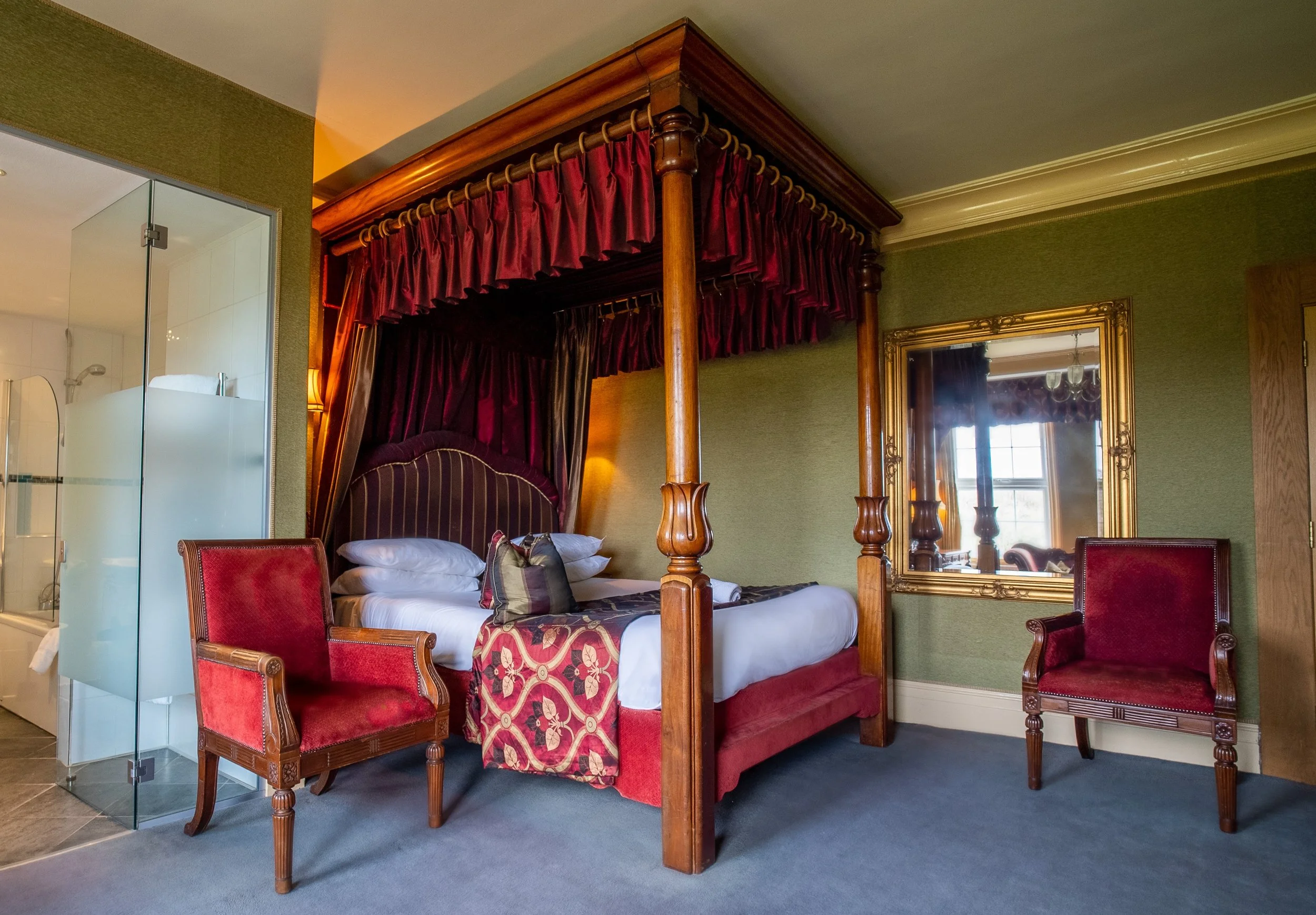 Hotel room with a four-poster bed, red velvet chairs, and a mirror on green walls.