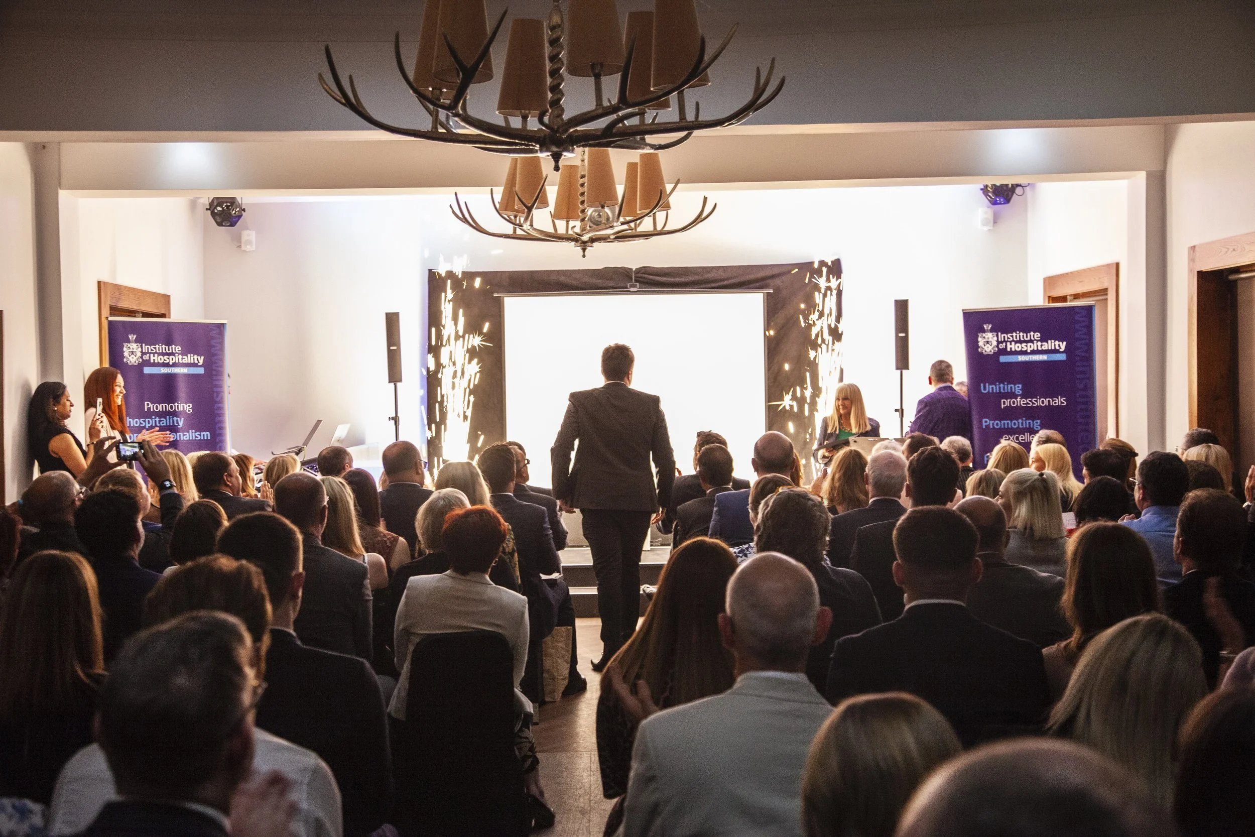 A large conference room filled with people attending a professional event. A presenter is walking towards the stage, which is highlighted by sparklers on either side. There are two banners with the logo of the Institute of Hospitality and the slogan 