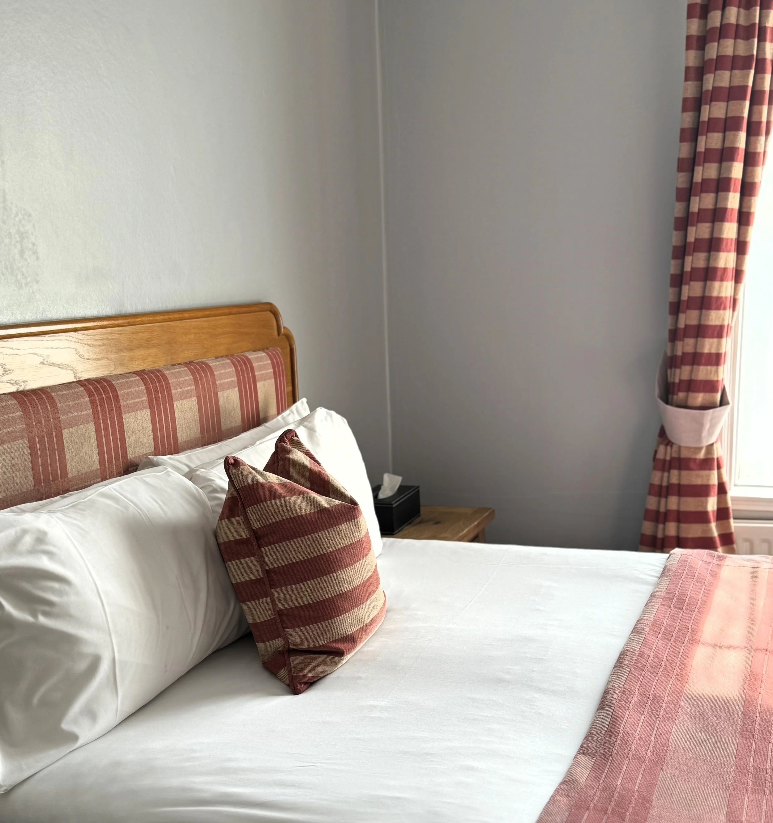 A neatly made hotel bed with white sheets, striped pillows, a matching runner at the foot of the bed, and a wooden headboard. There is a window with plaid curtains in the background.