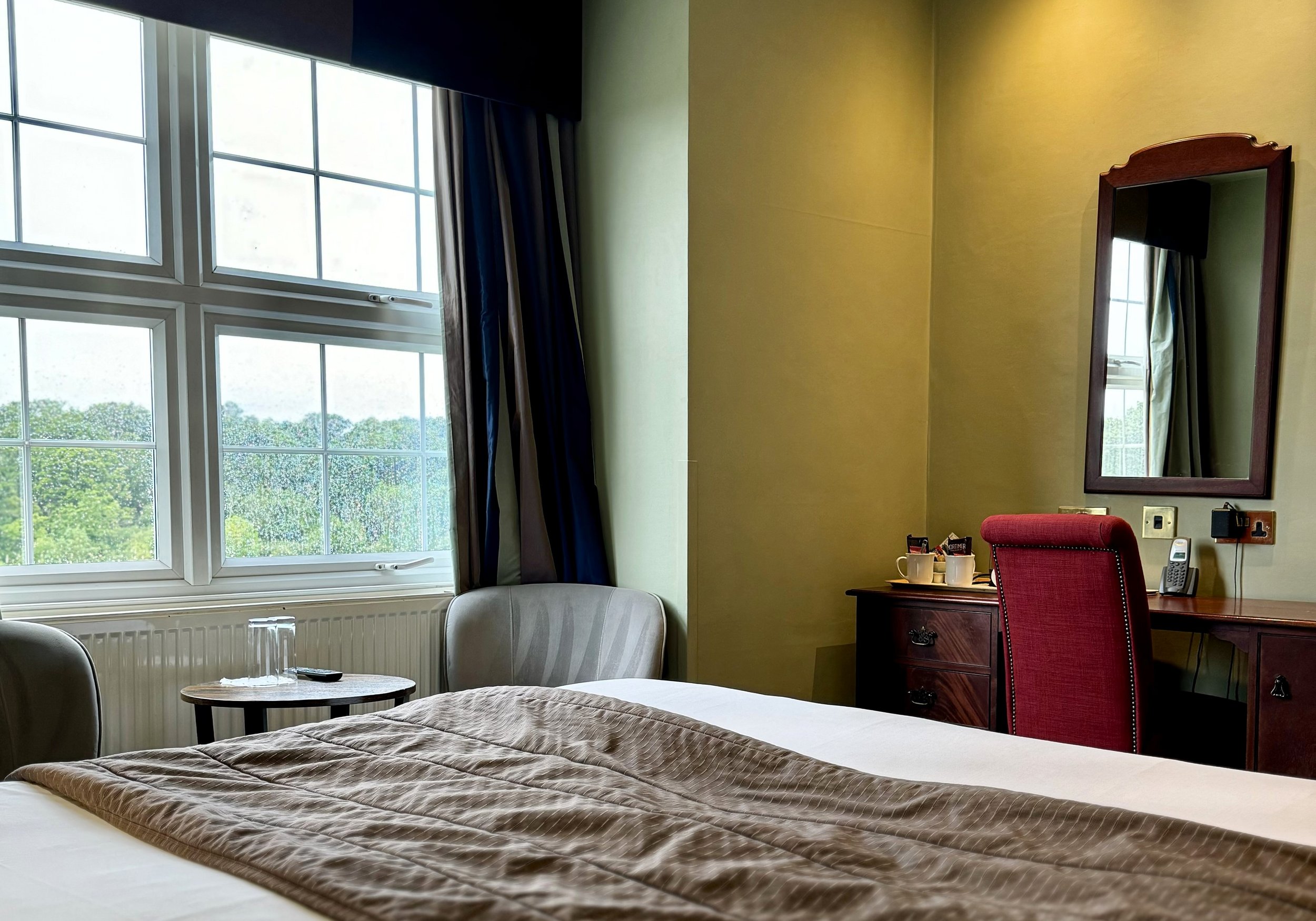 A cozy hotel room with a large window, a bed with a beige comforter, a side table with glasses and a remote, a dresser with cups, and a red chair in front of a mirror.