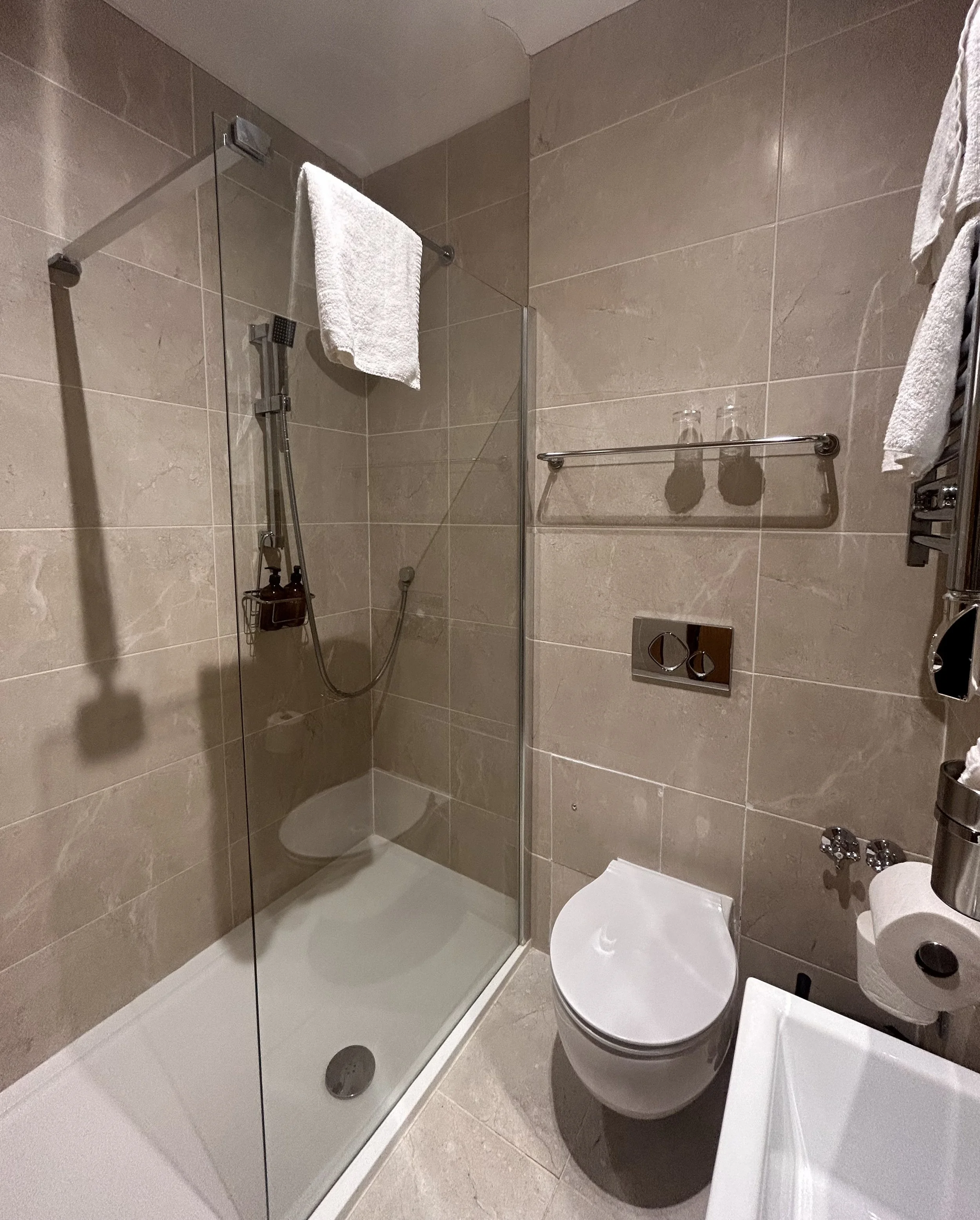 Bathroom with a glass shower enclosure, a wall-mounted toilet, a towel rack, and beige tiled walls and floor.