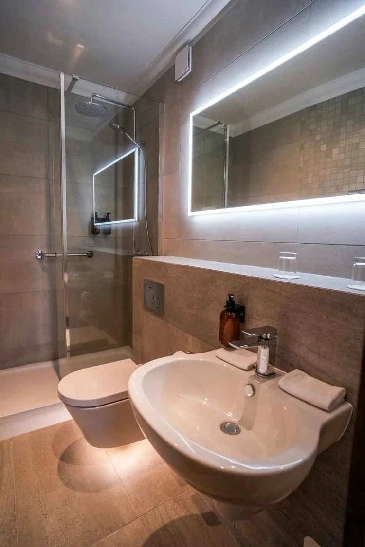 Modern bathroom with beige tiles, a walk-in shower with glass doors, a wall-mounted toilet, a white sink, and a large mirror with LED lighting.