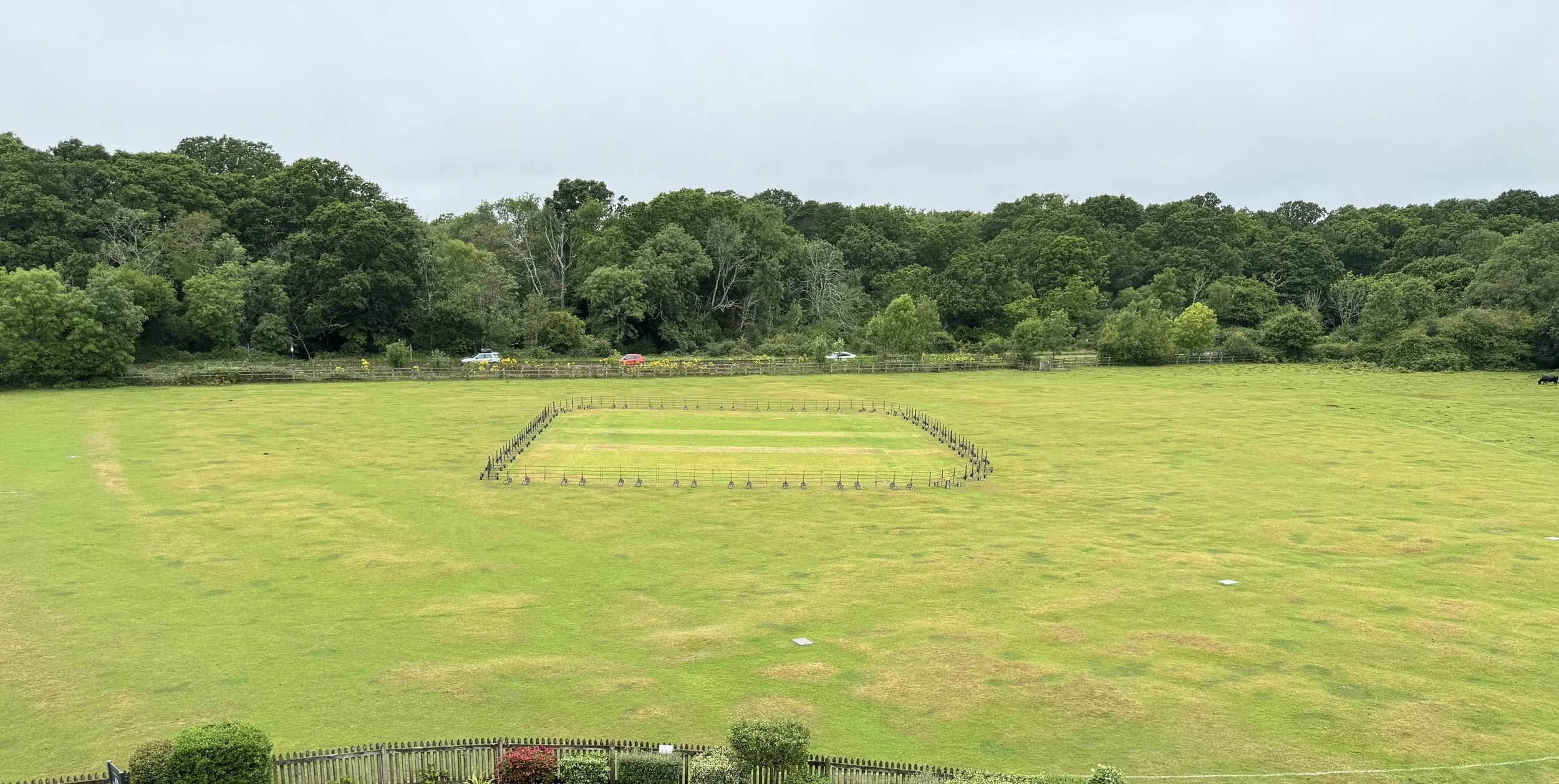 Large open grassy field with a rectangular fenced area in the center, surrounded by trees and a few cars in the background on a cloudy day.