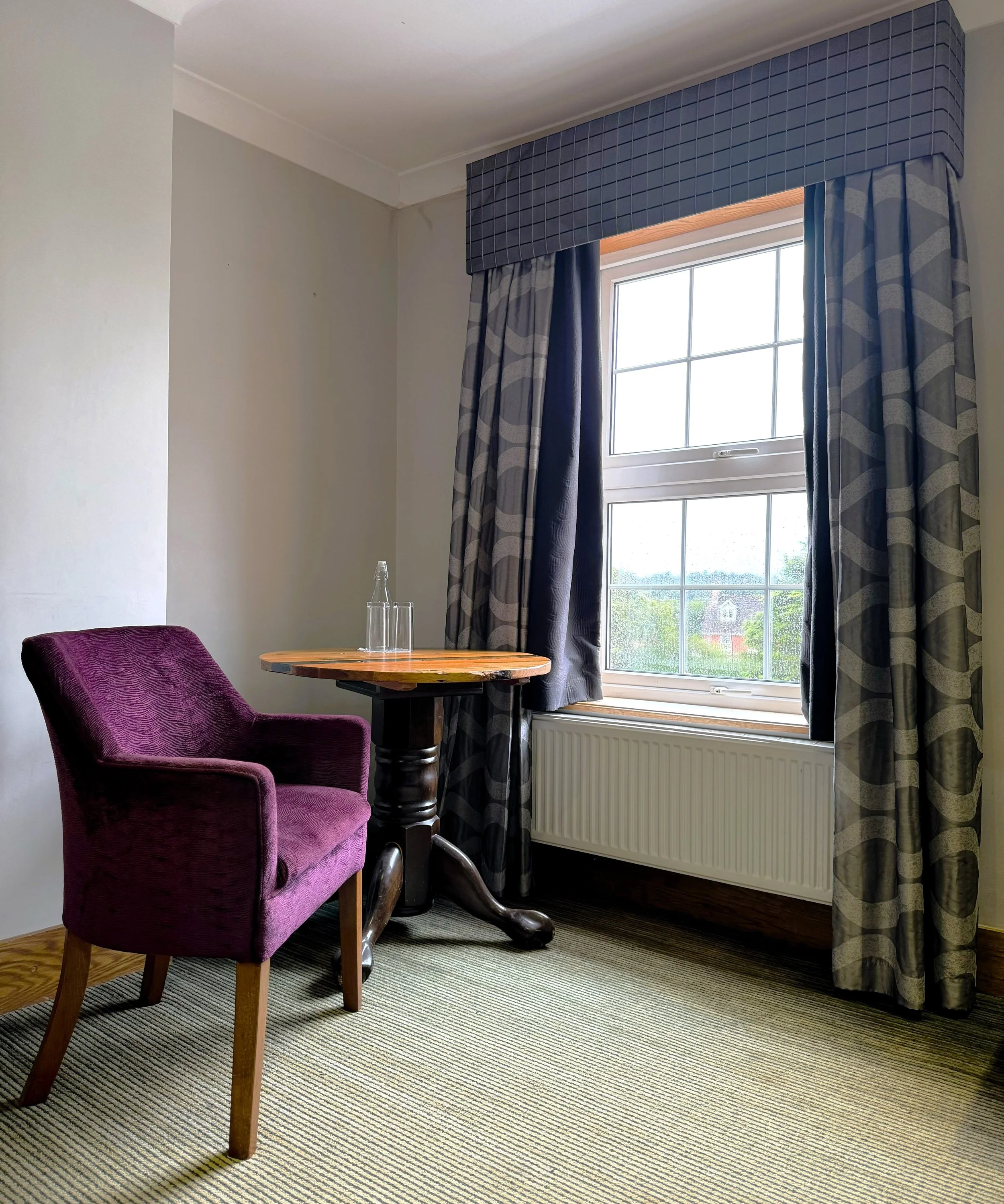 Interior of a room with a purple upholstered armchair, small wooden table, a glass bottle and glass, window with curtains, radiator, and a green outdoor scene visible through the window.